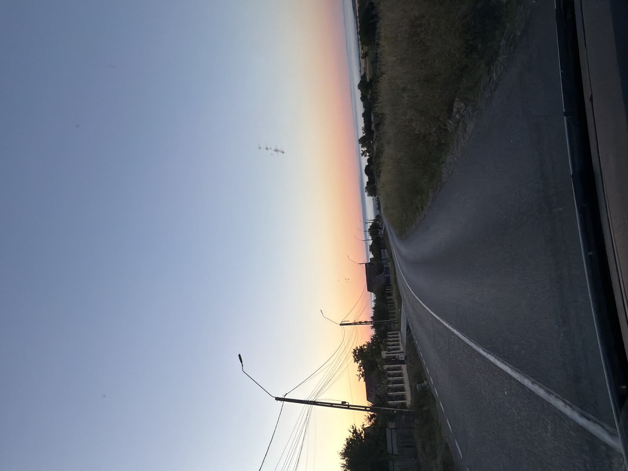 The image depicts a road in a rural area, possibly near Pașcani, Romania, during sunrise. The sky transitions from a pale blue at the top to hues of orange and pink near the horizon, indicating the early morning. A paved road curves through the scene, with a white line marking its center. On the left side of the road, there are houses with roofs and fences, along with utility poles and a complex network of wires. Tall streetlights are also visible along the roadside. The right side of the road is bordered by dry, tall grass and scattered trees. In the distance, a thin layer of fog or mist hovers over a body of water, possibly a river or lake. The image appears to be taken from inside a vehicle, as the bottom of the windshield is visible. There are no people or animals in the frame. The overall atmosphere is peaceful and serene, characteristic of a rural dawn.