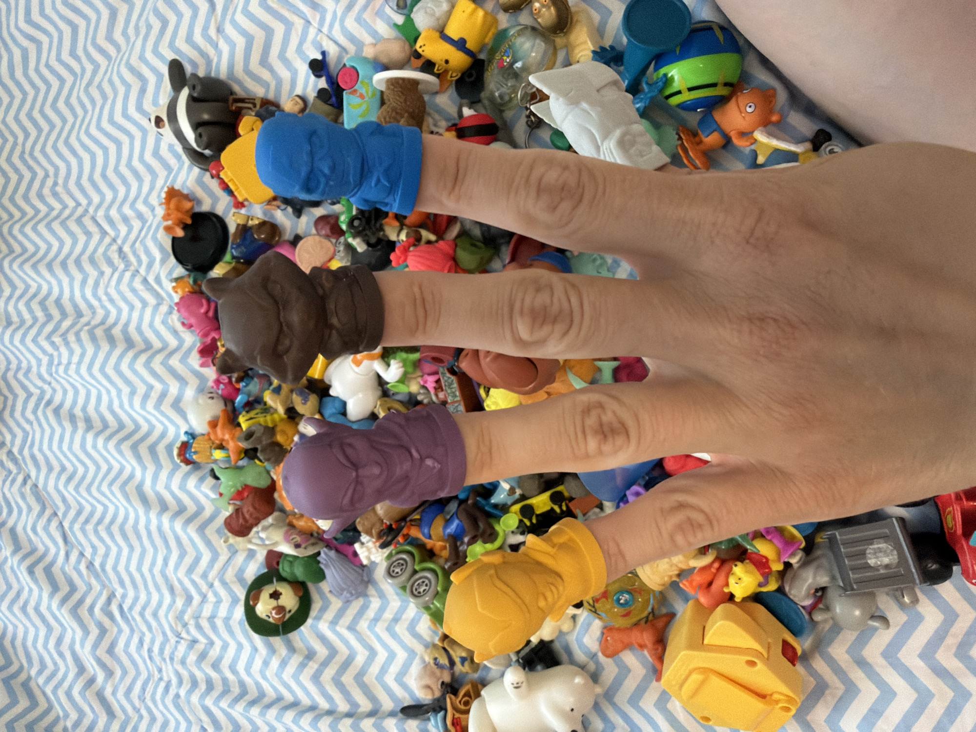 A person's hand rests on a pile of small toys scattered on a blue and white chevron-patterned surface, likely a bedspread or blanket. Each finger, excluding the thumb, is adorned with a finger puppet: blue, brown, purple, and yellow, respectively. The toys in the pile vary in color, shape, and apparent material, suggesting a collection of small figurines, miniature vehicles, and other miscellaneous playthings. The location is not discernible from the image and cannot be inferred to be Votkinsk, Russia, or any other specific location.