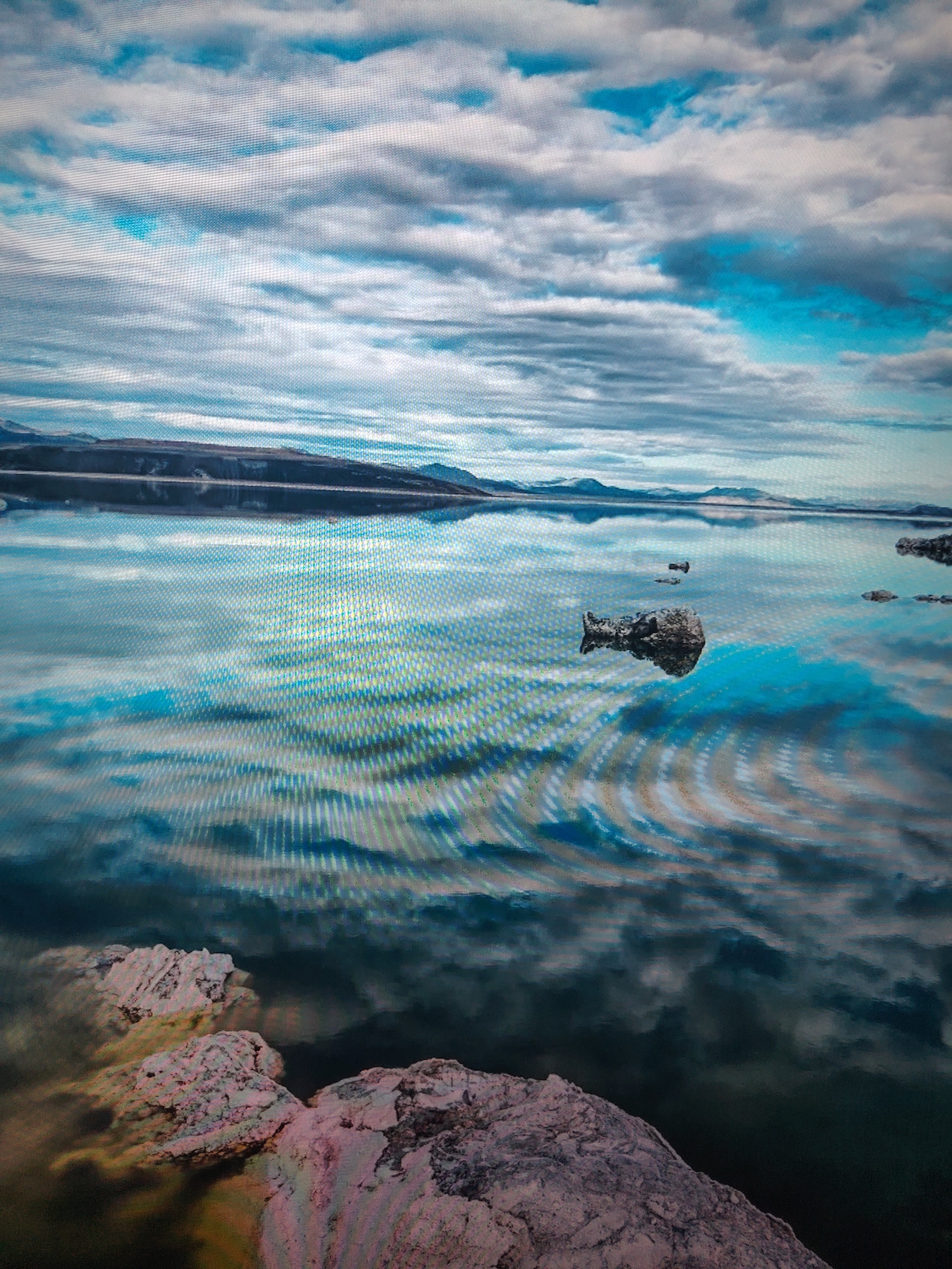 The image depicts a landscape with a body of water, possibly a lake, reflecting the sky. The sky is filled with clouds, ranging from white to gray, and patches of blue. Distant landmasses or mountains are visible on the horizon. The water's surface is rippled, creating distortions in the reflections. In the foreground, there are light-colored rocks with some pinkish hues. These rocks appear to be at the edge of the water or slightly submerged. Other smaller rocks or formations are seen protruding from the water further away. The image has a cool color tone due to the predominance of blues and grays. The location is Unknown.