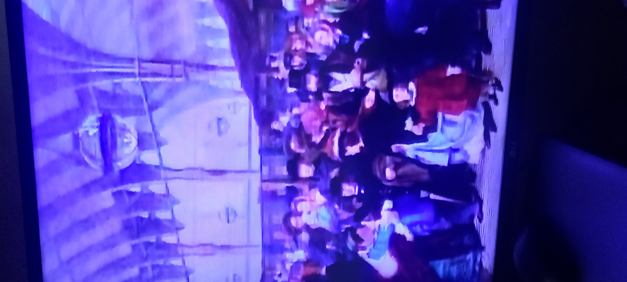 A dimly lit, dimly lit, and blurry, purple-tinted scene appears to show a gathering of people in what looks like a grand hall. The architecture above them suggests a medieval or gothic style, with arched ceilings and what might be hanging chandeliers. The people are dressed in what appear to be historical or costume attire, with many wearing hats and cloaks. The individuals are mostly standing, with some appearing to interact with each other. The scene is captured from a distance, and the overall quality of the image is poor, with a significant amount of blur and digital noise.