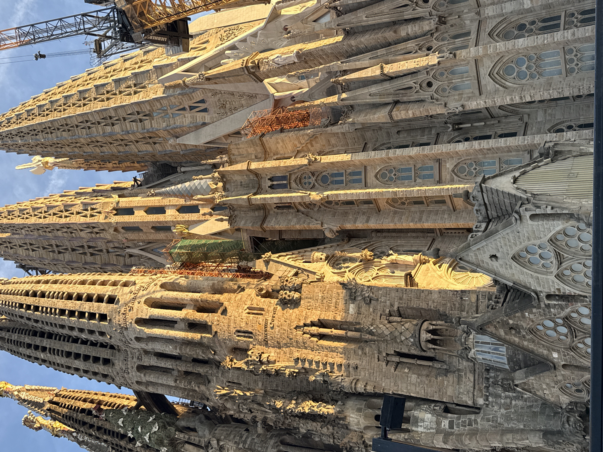 The image shows a close-up, low-angle view of the Sagrada Familia in Barcelona, Spain, bathed in the warm light of the setting sun. The iconic basilica's intricate facade, characterized by its stone towers adorned with countless openings and detailed carvings, dominates the frame. Cranes are visible in the upper right quadrant of the image, indicating ongoing construction or renovation work on the structure. The sky is a clear blue. The lower portion of the image displays sections of the basilica's lower architectural features, including pointed archways and decorative window designs.