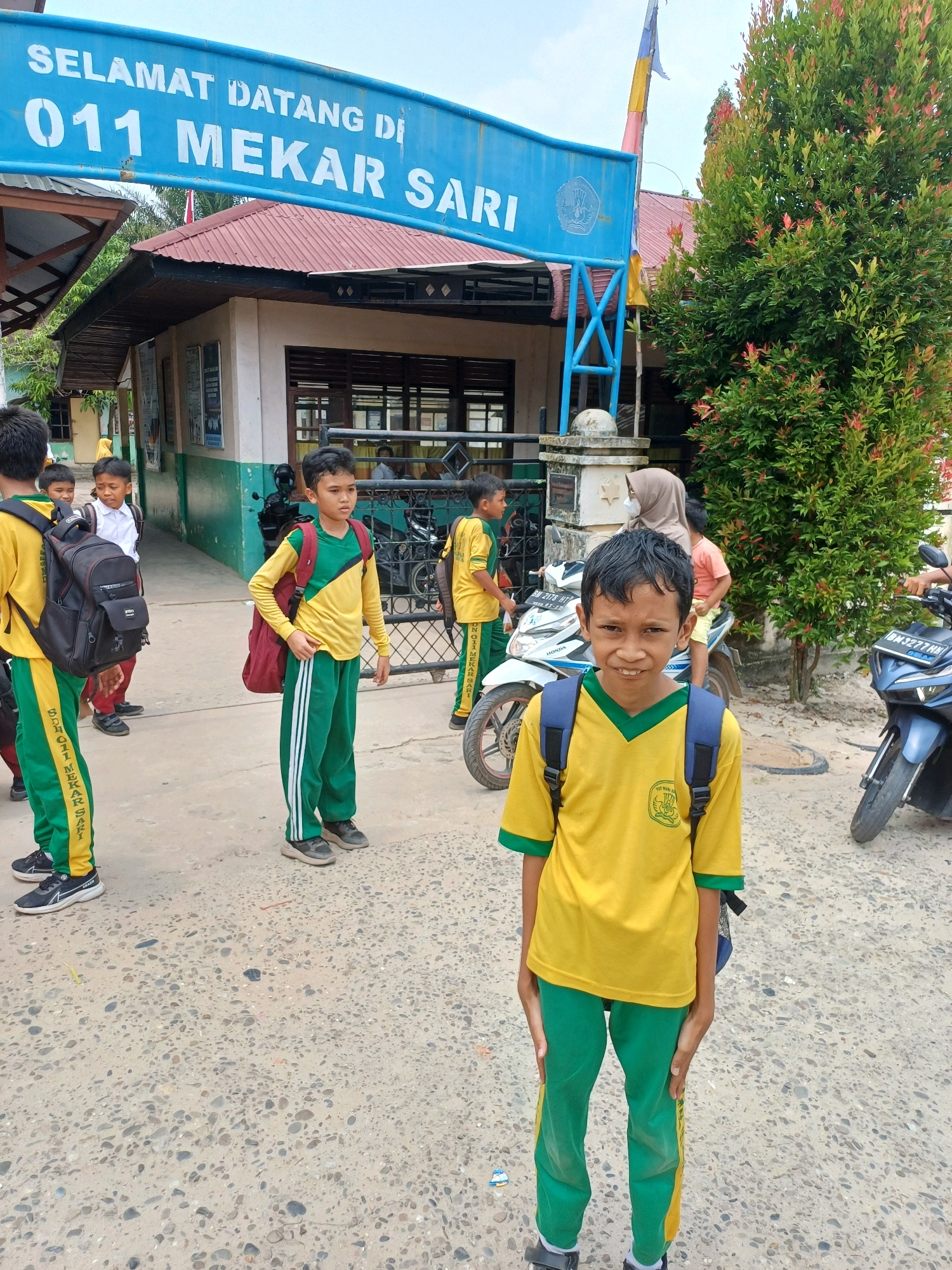 The image depicts the entrance of "SDN 011 MEKAR SARI" in Dumai City, Indonesia. A blue sign with white lettering welcomes visitors. In the foreground, a young boy wearing a yellow shirt with green trim and green pants with yellow stripes, carrying a blue backpack, smiles at the camera. Other children in similar school uniforms are visible in the background, some with backpacks and one on a white motorcycle. A blue motorcycle is parked to the right. The ground is paved with concrete and pebbles. Trees with red and green foliage are present on the right side of the frame. The sky is overcast.