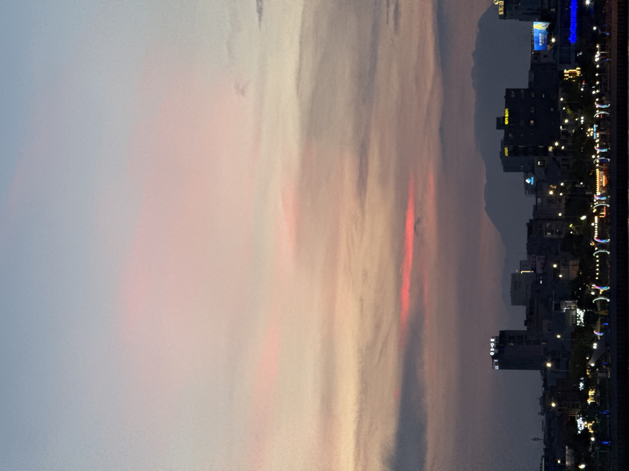 This is a landscape photograph taken at dusk in Da Nang, Vietnam. The image is oriented vertically, with the sky dominating the left side and a cityscape on the right. The sky exhibits a beautiful twilight palette, with soft hues of pink, orange, and pale blue blending together, suggesting the sun has recently set or is about to rise. Wispy clouds are visible, catching the last rays of light and creating streaks of color across the sky.
The right side of the image features a city skyline with several buildings of varying heights. Many of these buildings are illuminated by artificial lights, highlighting their structures and windows. The lights create a warm glow against the darkening sky. Along the waterfront, there are decorative lights, possibly part of a promenade or public space, featuring strings of colorful lights and curved illuminated structures.
Some visible text can be identified on the buildings. One building has "KIM SON" lit up in yellow. Another building has "BOND" visible in white illuminated letters. There is also text on a billboard in the background which is too blurry to read.
The overall mood of the image is serene and tranquil, capturing the transition from day to night in an urban setting. The combination of the soft sky colors and the vibrant city lights creates a visually appealing contrast. The weather appears to be clear, with no indication of rain. The presence of the cityscape and the well-lit waterfront suggests a populated and active urban environment.