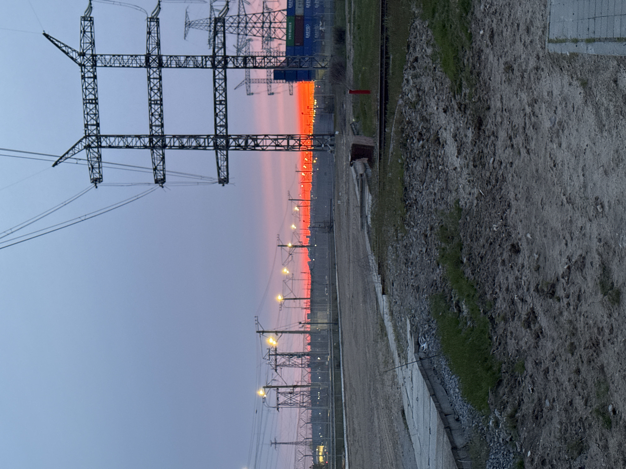 The image, taken in Volzhsky, Russia, depicts an expansive industrial or utility landscape at either dusk or dawn. The most striking feature is the sky, where the horizon glows with intense orange and red hues, gradually transitioning to shades of purple and light blue towards the top.
In the foreground, an unpaved path of dirt and gravel lies on the right, interspersed with patches of green grass. A concrete pathway runs parallel to it on the left. The midground is characterized by a long, straight stretch, possibly a railway track or an industrial road, which recedes into the distance. It is lined with numerous utility poles bearing streetlights that emit a warm, yellow illumination, their lights forming a distinct line against the colorful horizon. Large, lattice-structured transmission pylons and intricate power lines are prominent features, silhouetted against the vibrant sky.
In the upper left portion of the image, stacked shipping containers are visible, further emphasizing the industrial setting. Text such as "EVERGREEN" and "EASTOP" can be discerned on some of these containers. The scene is devoid of any visible human activity, conveying a sense of vastness and quiet contemplation of the industrial infrastructure under a dramatic twilight sky.