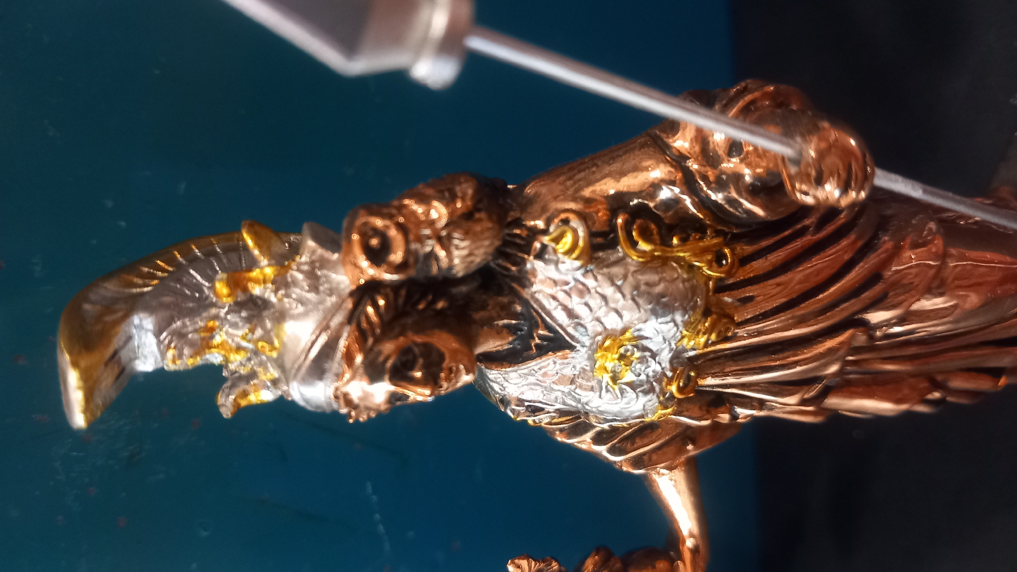 The image is a close-up, eye-level shot of a metallic figurine of Athena, the Greek goddess of wisdom and warfare. The figurine is predominantly bronze with silver and gold accents. Athena is depicted wearing a helmet with a crest, ornate armor, and a flowing robe. An owl, a symbol of Athena, is perched on her left shoulder. She holds a spear in her right hand and what appears to be a small object, possibly a Nike figurine, in her left hand. The background is a mottled teal blue surface. The lighting highlights the metallic sheen of the figurine. The overall impression is of a detailed and decorative artifact. The image does not provide any information about the city of Kos, Greece.