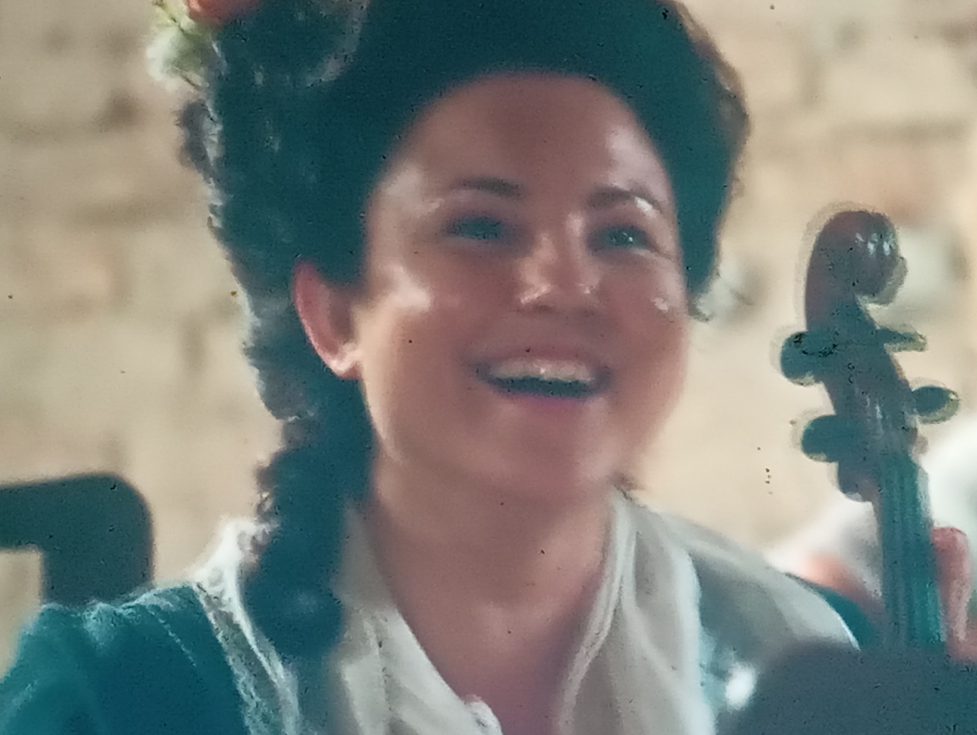 A close-up, slightly blurred, and grainy shot shows a woman with dark hair styled in an updo, adorned with flowers, and a braid hanging down her back. She is wearing a teal jacket with a white ruffled collar and appears to be smiling broadly, revealing her teeth. Her eyes are looking upwards and to the right. To the right of the woman, a dark, blurry object with curves suggests a musical instrument, possibly the scroll and tuning pegs of a cello or violin. The background is a soft, out-of-focus beige wall, possibly brick or plaster, with some indistinct shapes of other people or objects. The lighting is soft, and the overall impression is of a candid moment, perhaps during a performance or celebration, with a joyful and lively emotion conveyed by the woman's expression. There is no visible text in the image.