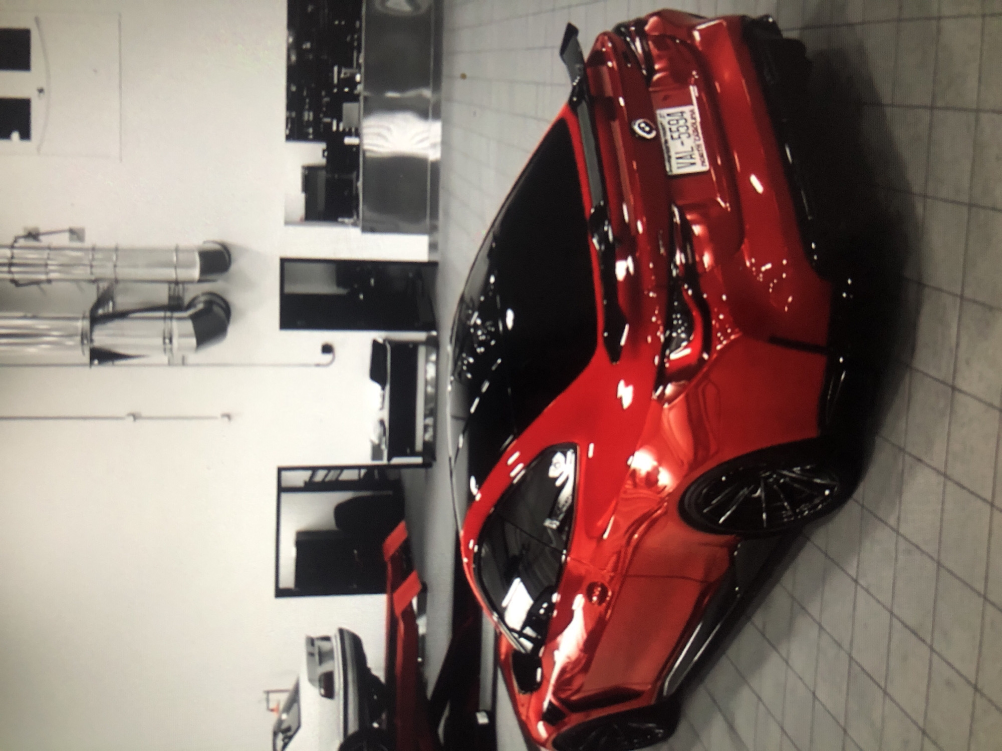 The image shows a red BMW M4 coupe parked in what appears to be a car shop, with a white BMW sedan and a red BMW sedan also visible in the background. The M4 coupe has a dark tinted roof and a North Carolina license plate with the number VAL-5594. The environment is a garage or shop with a white tiled floor and light grey or white walls. The location in Lhokseumawe, Indonesia cannot be determined from the image.