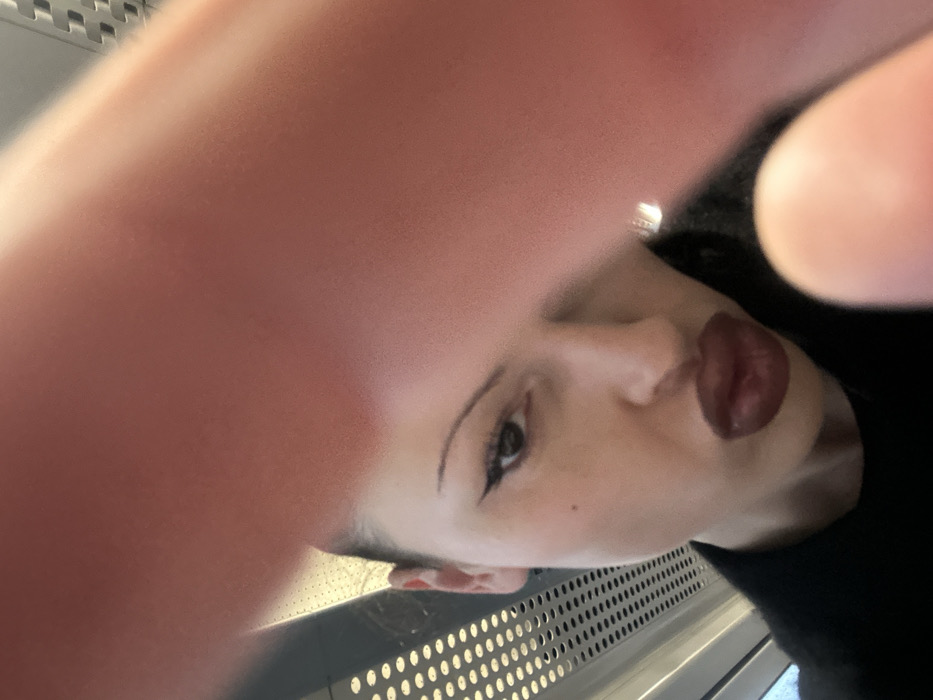 The image depicts a young adult female in a close-up, angled selfie taken within a train interior. The subject's head is slightly tilted, with her eyes fixed on the camera and her lips pursed. Her facial features include dark, pulled-back hair, drawn-on arched eyebrows, pronounced winged eyeliner, and dark, over-lined lips. A small pigmented lesion is present on her left cheek. She is dressed in a dark garment. The upper and lower right foreground is blurred by a large, out-of-focus, light-skinned digit, likely a finger or thumb, partially obscuring the subject's face. The background on the left reveals a metallic panel with a horizontal grid of circular perforations, characteristic of a modern train carriage interior. This setting is consistent with public transportation that would operate through or to Sevenoaks, United Kingdom; however, no specific landmarks or unique identifiers are present within the frame to establish a precise location such as a station or street within Sevenoaks.