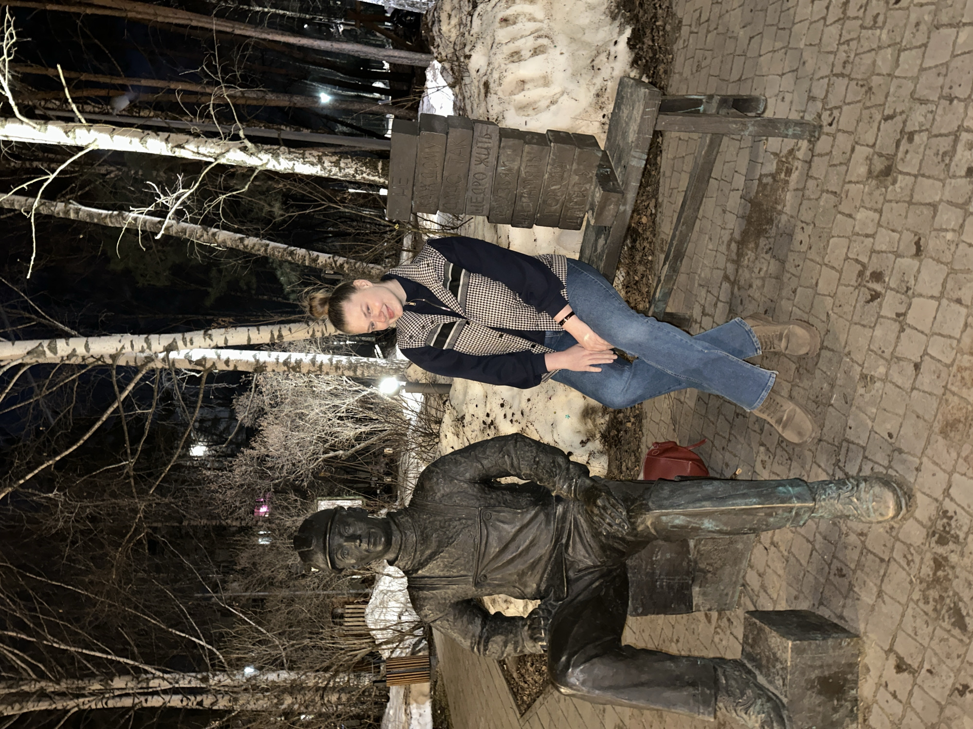 A woman is posing with a bronze statue in Kazan, Russia. The woman, positioned above the statue, wears a black and white houndstooth jacket, blue jeans, and tan boots. The statue depicts a person in work attire, seated, with a cap on. Behind the woman is a wooden bench, and stacked blocks of stone with inscriptions in Russian. The surrounding environment includes bare trees and a ground paved with stone blocks. Patches of snow are visible near the bench.