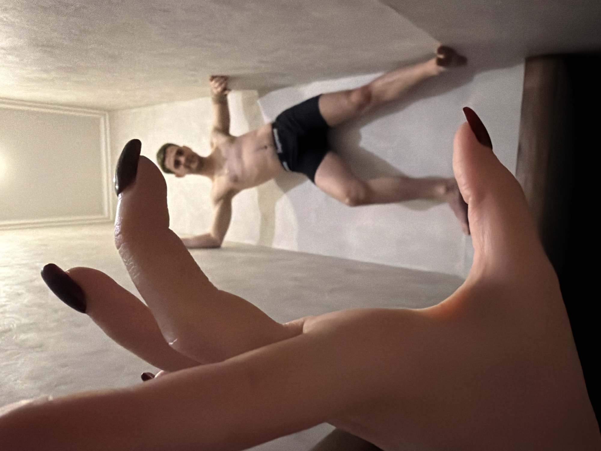The image shows a man lying on what appears to be a wall, clad in black boxer briefs, with his legs extended to the right and his arms outstretched. The background reveals a room with a plain ceiling and a framed section on the wall. In the foreground, a hand with dark-colored nails is prominently displayed, seemingly held up in front of the lens. The scene's location is unspecified. The interactions and actions involve a person posed on the wall and another person holding a hand towards the camera. The environment looks to be an indoor space.