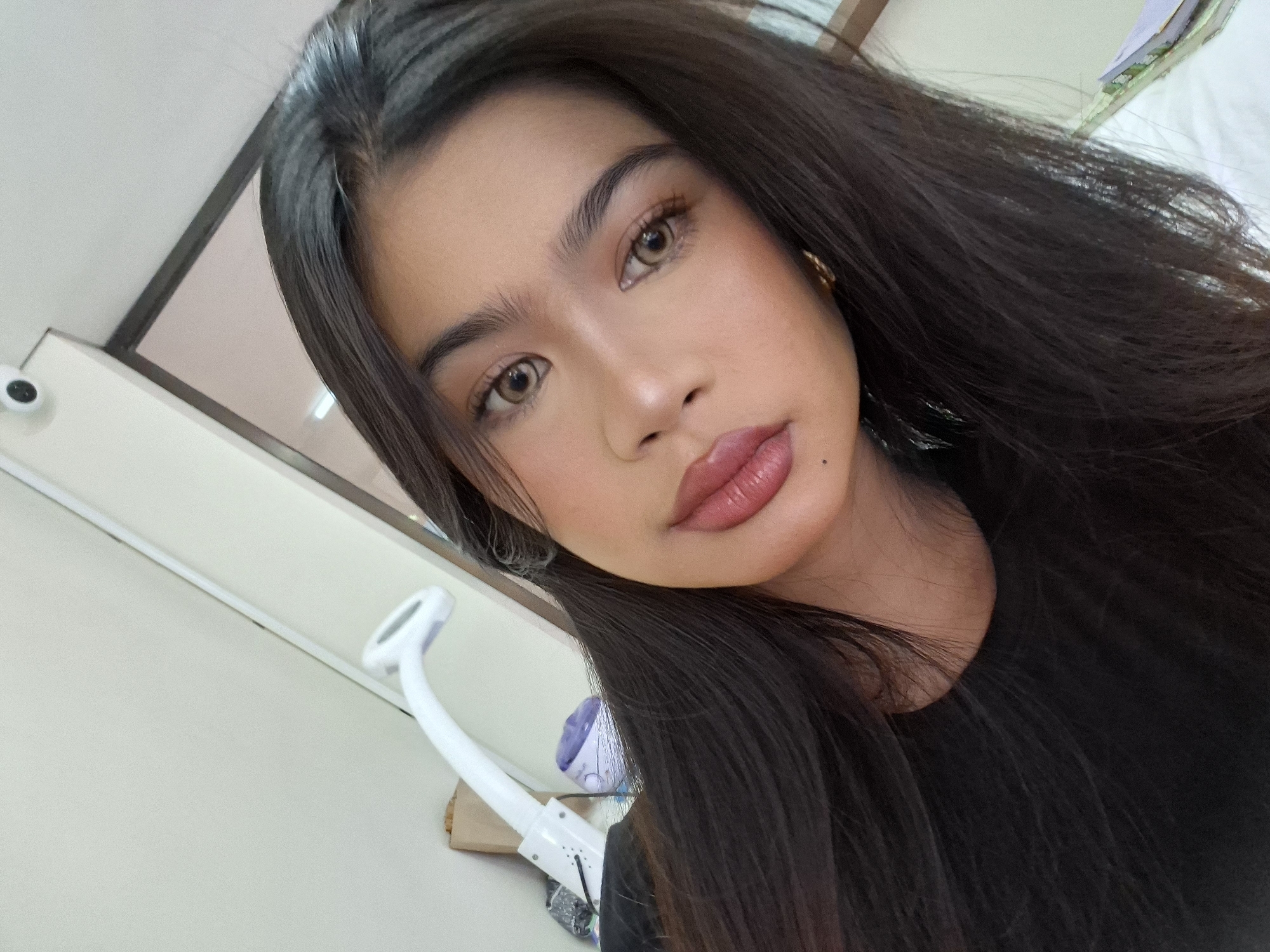 A selfie of a young woman with long, dark hair and light brown eyes is shown. She is wearing a black top and has on makeup, including lipstick and eye makeup. In the background, a white lamp with a flexible arm is visible, along with a brown paper bag and a small white device that appears to be a security camera. The setting suggests an indoor environment, possibly a room or studio. The image is taken from a close-up perspective, focusing on the woman's face. There is no indication of the image being taken in Kuta, Indonesia.