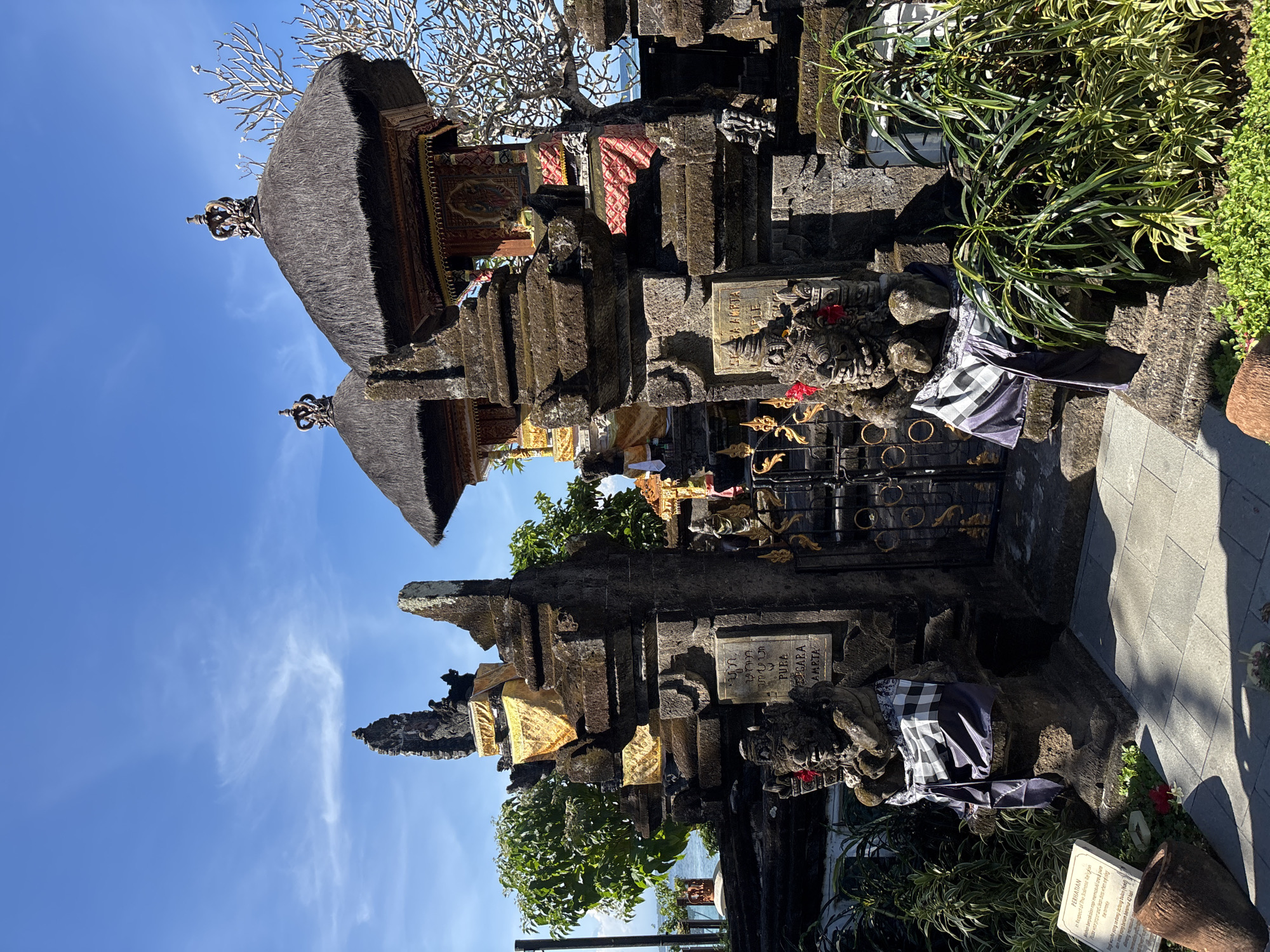 The image shows the Pura Segara Amerta temple in Bali, Indonesia. The temple is constructed of dark stone and features traditional Balinese architecture, including thatched roofs and intricate carvings. Strips of colorful cloth and patterned sarongs adorn the temple structures. Plants and greenery surround the temple, and the sky is blue with wisps of clouds. Signs with Balinese script and English text are visible, providing instructions for visitors, such as dress code and restrictions for menstruating women. The overall impression is that of a sacred and well-maintained cultural site.