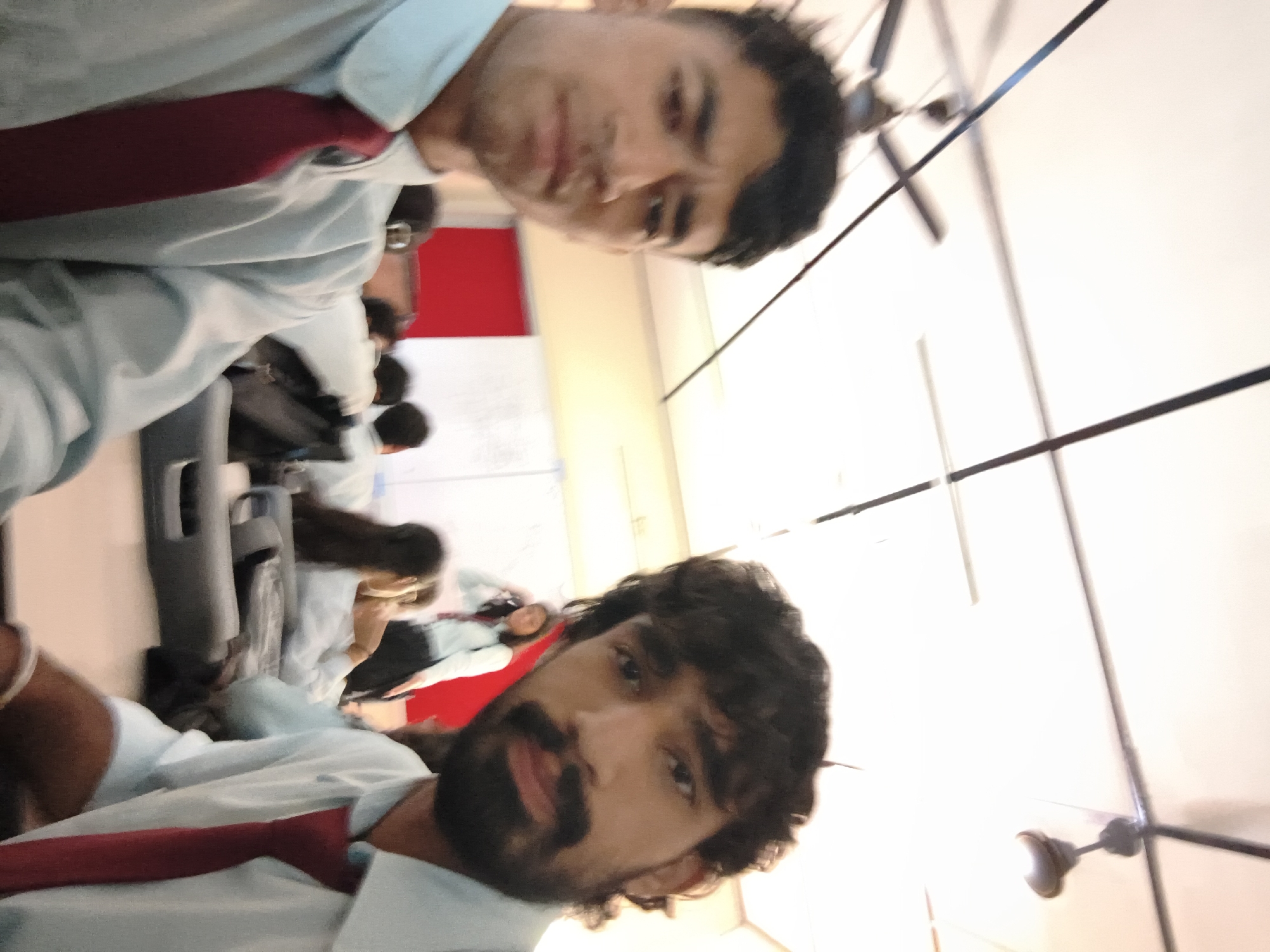 In a classroom setting in Dharamshala, India, two young men, presumably students, are taking a selfie. The foreground is dominated by their faces and upper bodies, both wearing light blue shirts and red ties, suggesting a school uniform. The man on the left has short, dark hair and a somewhat furrowed brow, while the man on the right has longer, curly hair and a beard. Behind them, other students are visible, some seated at desks and others standing. One female student in the background wears a similar light blue shirt and dark pants. The classroom appears to have whiteboards on the walls, and ceiling fans are present, indicating a typical educational environment. A bright red panel is visible on the wall behind the students. The lighting suggests it is daytime, possibly with fluorescent lights contributing to the illumination.