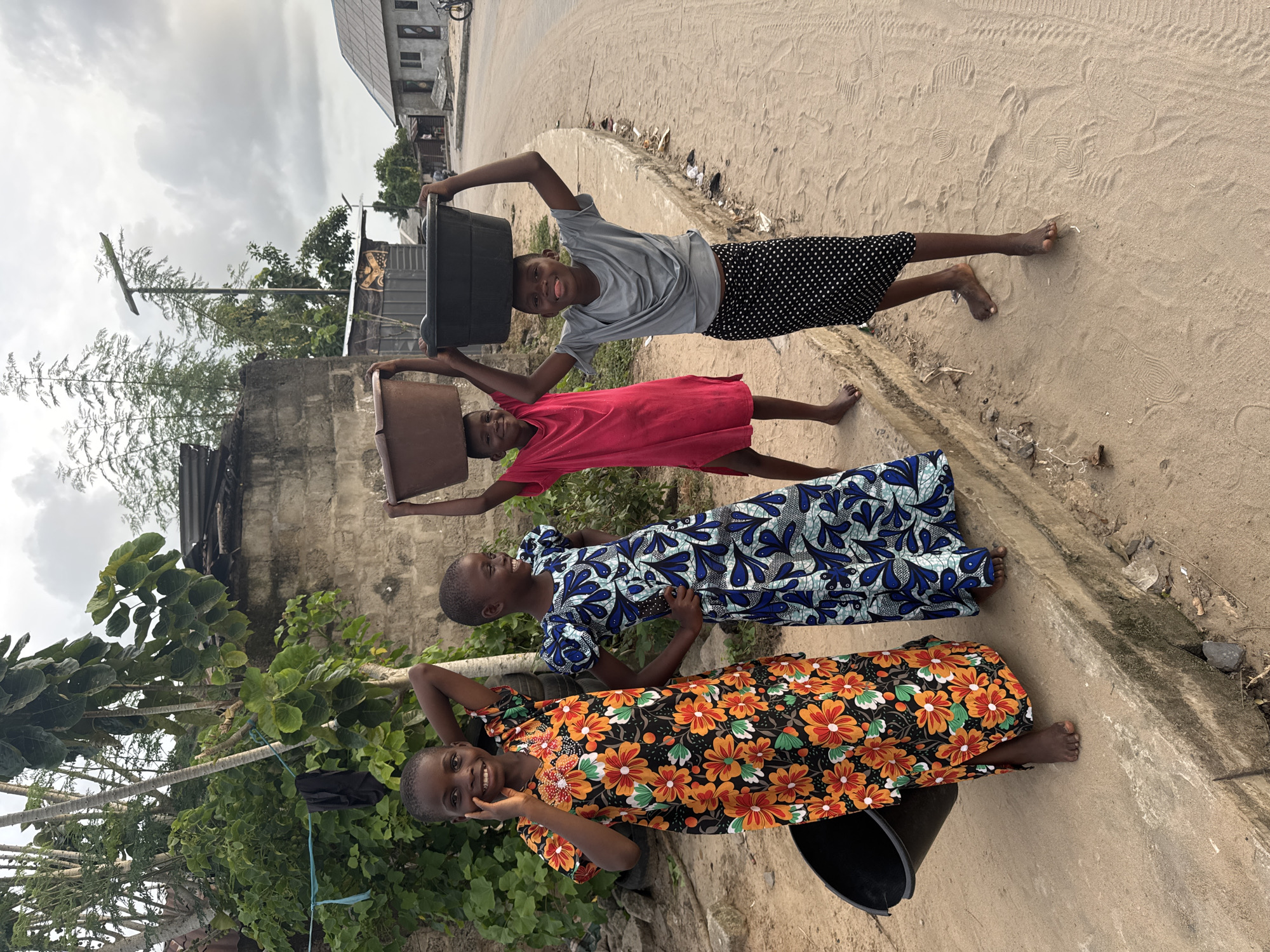 Four young girls, presumably sisters, stand barefoot on a sandy area beside a paved road in Badagry, Nigeria. Each balances a container on their head. Starting from the front, the first girl wears a black dress with orange and yellow floral patterns and holds a black bucket. The second girl wears a blue and white patterned dress. The third girl is in a red dress. The last girl wears a grey shirt and black polka dot shorts. They are all smiling and facing forward. Behind them are buildings with corrugated iron roofs and a concrete wall. Vegetation is also visible. The sky is cloudy.