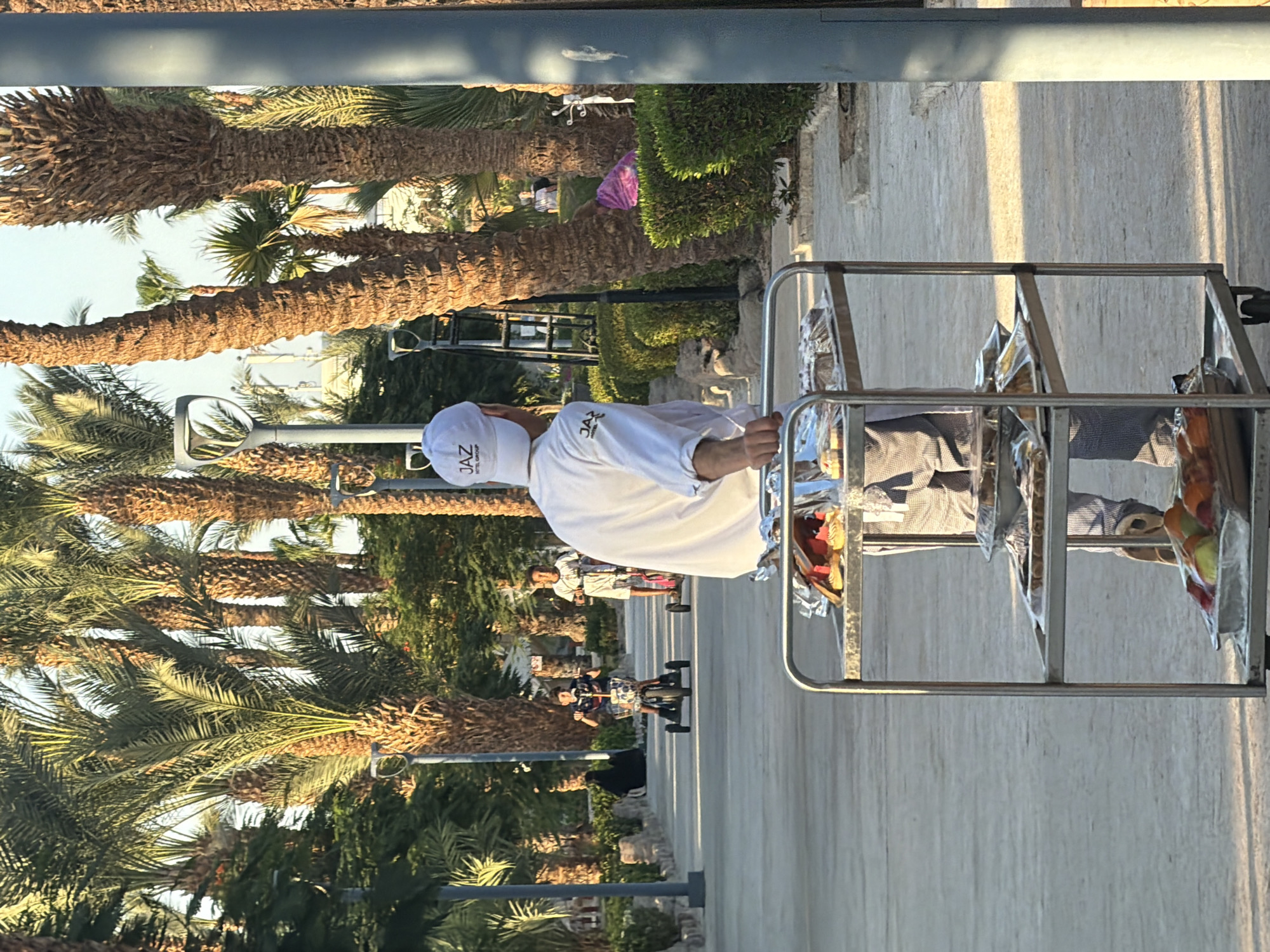 An outdoor scene in Sharm El Sheikh, Egypt, features a person in a white chef's uniform and a white baseball cap with "JAZ HOTEL GROUP" on it, pushing a food cart. The cart has three shelves laden with food in foil-covered trays and plastic packaging, including what appears to be pastries and fruit. The person is standing on a paved walkway lined with palm trees and green bushes. In the background, two young boys are riding small electric scooters on the walkway. Another person, wearing a black abaya, is seated on a stone bench to the left, and a partially visible person in a pink garment is in the background to the right. The lighting suggests it is late afternoon or early morning, with long shadows cast by the palm trees.