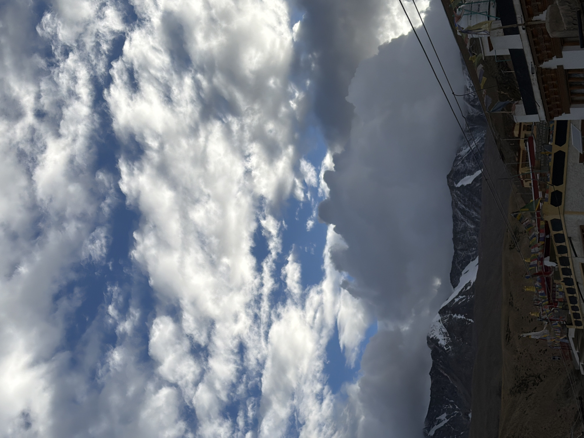 The image depicts a dramatic sky with a mix of dark, heavy clouds and bright, illuminated clouds, set against a backdrop of snow-capped mountains. In the foreground, a portion of buildings in Karzok, India, is visible. These buildings feature traditional Tibetan architecture with white walls, dark trim, and ornate detailing. Prayer flags are strung across the rooftops, adding a splash of color to the scene. The overall impression is one of a rugged, high-altitude landscape under a dynamic and imposing sky.