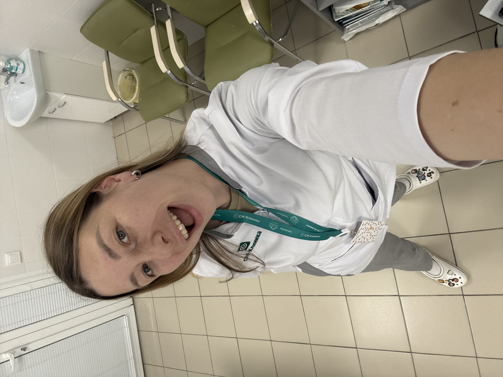 A young Caucasian woman, appearing to be in her late twenties or early thirties, is taking a selfie in what appears to be a medical clinic waiting area. She has fair skin with freckles across her nose and cheeks, blue eyes, and light brown hair. She is wearing a white medical jacket over a tan t-shirt, gray pants, and white Croc-style shoes adorned with various charms. A green lanyard with a medical clinic logo is around her neck, and a badge is clipped to her jacket. Her mouth is open in a wide smile, and she is looking directly at the camera.
The environment is a tiled room with light-colored walls and floor. To the left, a white window blinds are visible behind a frosted glass door. In the background, there are two green chairs with chrome frames, a white sink with a chrome faucet and soap dispenser, and a small white cabinet. A metal trash can with yellow contents is situated between the chairs. To the right, a shelf unit holds various medical supplies and documents. The overall impression is a clean, functional space within a medical facility.