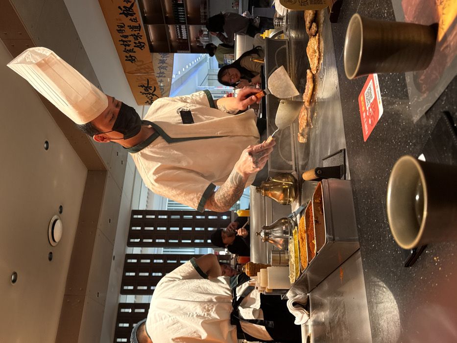Within a teppanyaki restaurant in Liangjiang, China, a male chef is observed cooking various cuts of meat on a large flat-top grill. The chef, positioned centrally, wears a tall white paper chef's hat, a black face mask, and a white uniform with green trim, revealing tattooed forearms. He is actively manipulating the meat with two metal spatulas, generating steam visible above the grill surface. To his immediate left, the back of another chef, wearing a white uniform and black apron, is partially visible.
In the foreground, two dark, cylindrical cups containing liquid are placed on a speckled black countertop, alongside a red square sticker displaying a QR code. Behind the cooking area, a metal tray filled with various powdered spices (including red, orange, and yellow hues) is present, alongside silver and brass-colored cooking vessels.
The background features several seated patrons. One female customer with glasses observes the cooking from the mid-right. Further back, a male customer in a dark hat holds an item to his mouth. Decorative wooden screens with vertical slats divide sections of the restaurant. Above, banners with Chinese characters are suspended, and shelves stocked with bottles are visible. The ceiling is equipped with overhead recessed lighting and circular air vents, illuminating the indoor environment.
