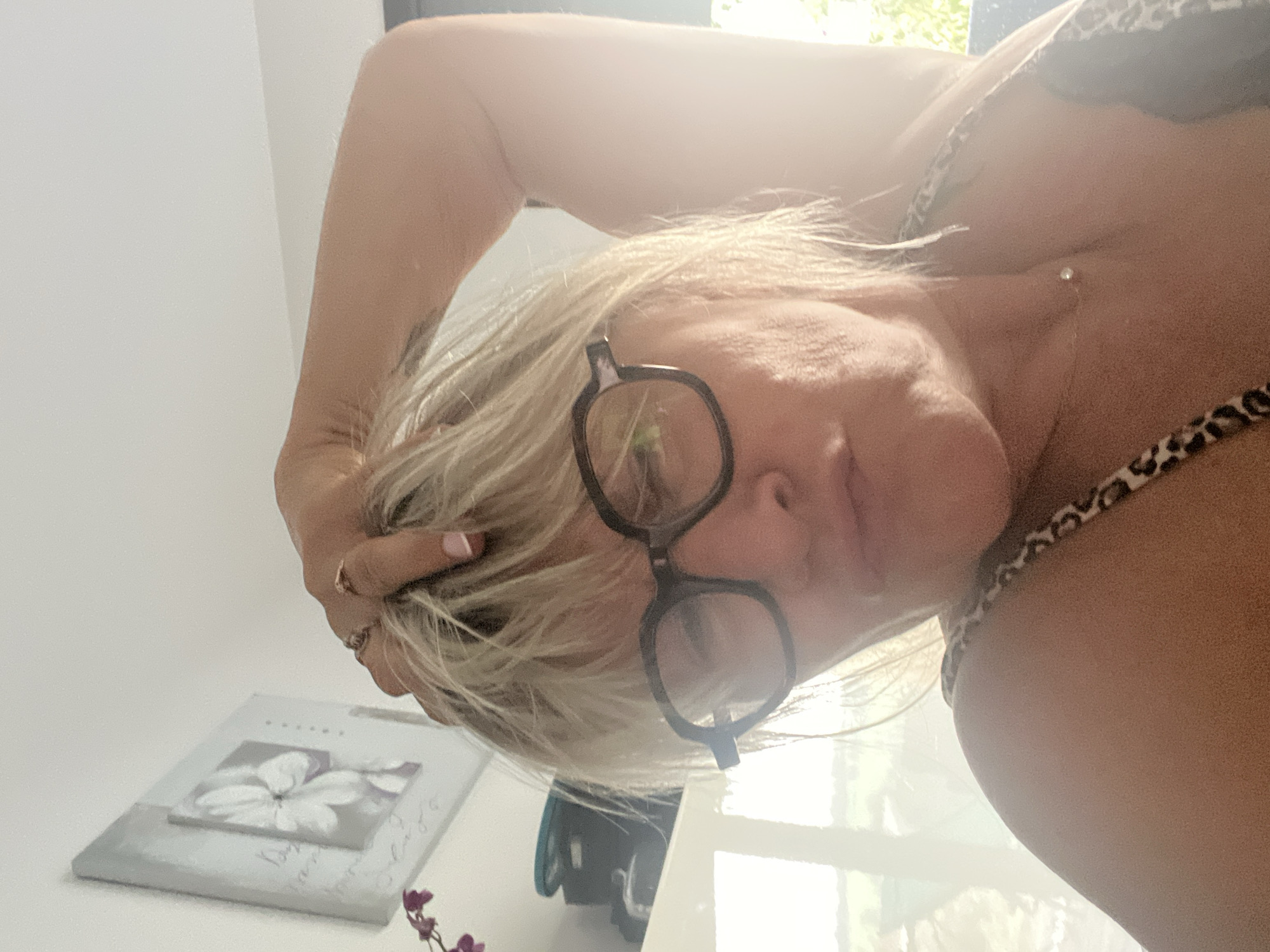 In Chambœuf, France, a woman with blonde hair and glasses is captured in a close-up selfie. She is wearing a leopard print top and a delicate necklace with a single pendant. Her right arm is raised, with her hand resting on top of her head, and her fingers are adorned with rings. The background features a framed picture of a white flower on the wall and a bright window with foliage visible outside. The lighting suggests an indoor setting with natural light streaming in.
