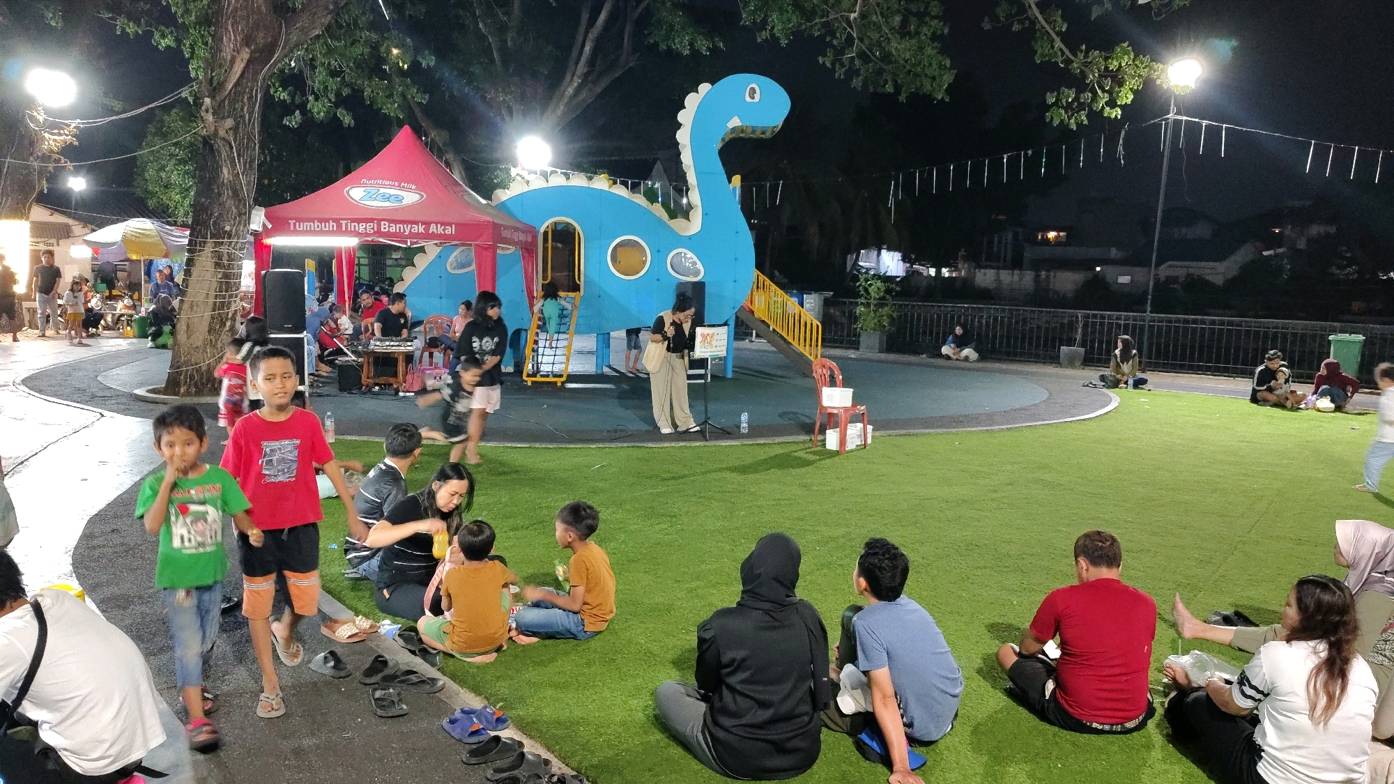 This is a nighttime photograph of an outdoor gathering in the city of Tangerang, Indonesia, likely at a playground or park area. A large, blue dinosaur-shaped play structure is prominent in the background, with yellow slides and stairs. To the left of the dinosaur structure is a red tent with a sign that reads "Tumbuh Tinggi Banyak Akal" and "Nutritious Milk". Several people are gathered, with some sitting on the green artificial turf in the foreground and others standing or walking around.
In the foreground, two young boys are walking towards the camera. The boy on the left wears a green t-shirt and blue jeans, while the boy on the right wears a red t-shirt and black shorts with orange accents. Several adults and children are seated on the grass, some facing a woman standing near the dinosaur structure with a microphone. A white speaker is also visible near the tent. In the background, beyond the play area, there is a fence and some buildings, illuminated by streetlights and string lights. Various food stalls or smaller tents are visible to the far left. The overall scene suggests a community event or a casual evening outing.