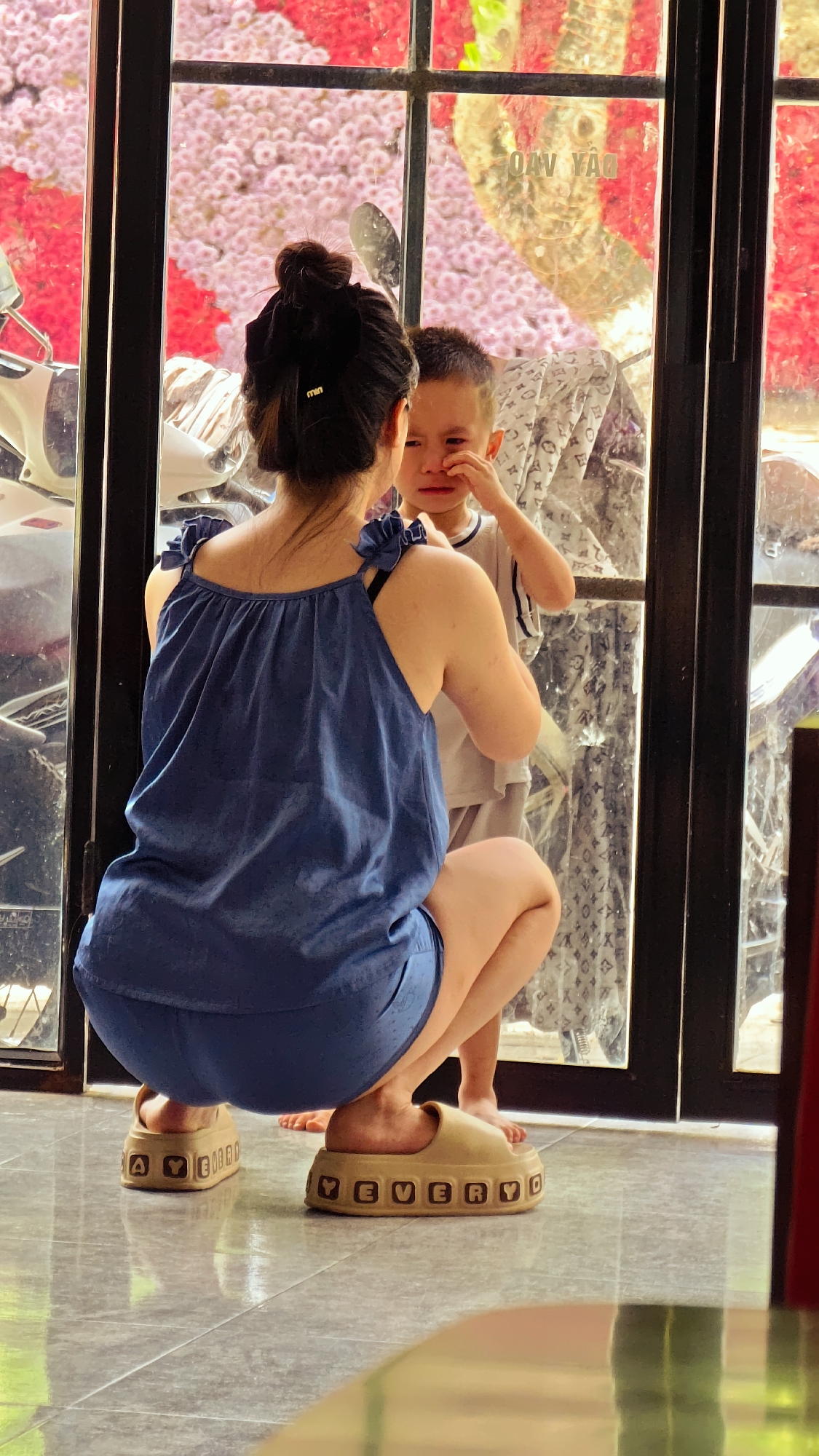 An adult female and a young male child are situated indoors on a light-colored tiled floor in Trung Tam Ward, Vietnam, positioned in front of a large glass window with dark frames. The woman, viewed from behind, wears a navy blue sleeveless top with ruffled straps, revealing a black bra strap, and matching navy blue shorts. Her dark hair is styled in a bun, secured with a dark ribbon and a small golden "MIN" hair clip. She is in a squatting position. On her feet are light brown, chunky platform slide sandals with raised letters spelling "AYE EVE" on the left and "EVERYD" on the right, indicating "EVERYDAY" or similar. Her bare feet are visible within the sandals.
The young male child, partially obscured by the woman, faces her with a distressed expression, crying and rubbing his left eye with his left hand. He wears a light-colored t-shirt with dark trim on the collar. Through the window, vibrant pink and red flowers or foliage are visible in the background, along with a partial view of a white scooter on the left. Reflections on the glass include a faint tree trunk, patterned fabric (possibly with a logo), and reversed text "OAV YAQ."