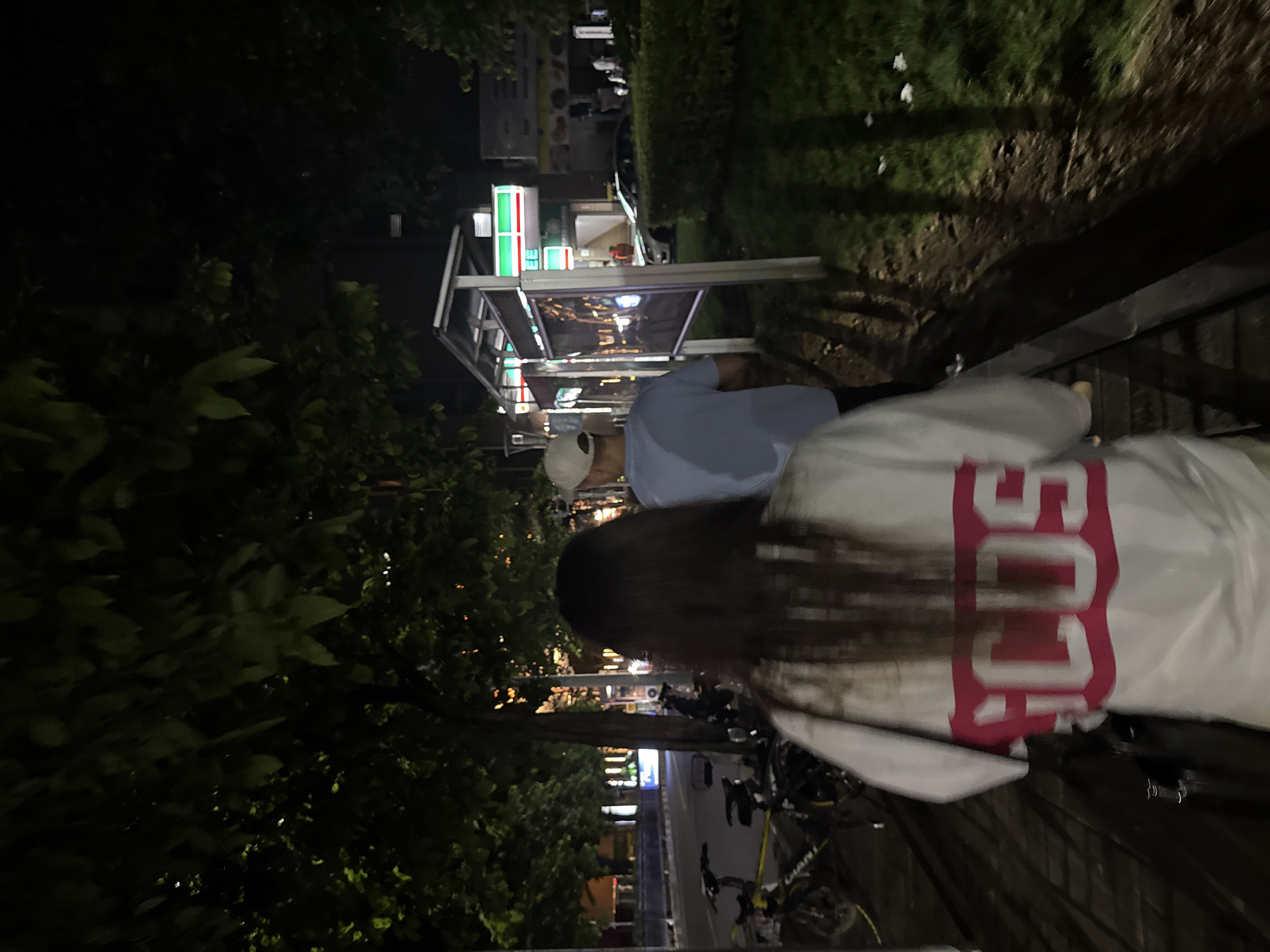 The image depicts a nighttime scene on a street in Haizhu District, China. The primary subjects are two individuals, partially obscured, and a street-side establishment with illuminated signage.
One individual, facing away from the camera, is wearing a light blue t-shirt and a white cap. They appear to be standing or walking. The other individual, also facing away, has long dark hair and is wearing a white jacket or shirt with prominent pink lettering that reads "CCDS".
The setting appears to be a commercial area at night. A structure with a canopy and illuminated panels is visible, possibly a storefront or a food stall. The signage on this structure includes the iconic red, green, and white logo of "7-Eleven". Beyond this, other businesses with glowing signs are discernible, suggesting a lively commercial street. There are also trees with dark foliage lining the path. In the foreground, a wooden bench or railing is present, and bicycles are parked nearby, indicating a public or pedestrian area.
The time of day is clearly night, evidenced by the artificial lighting and the darkness of the surroundings. The weather appears to be calm, with no strong indications of rain or wind. The overall mood is casual and urban, typical of a street scene at night in a bustling district. The visible text includes the "7-Eleven" logo and the "CCDS" lettering on the jacket.