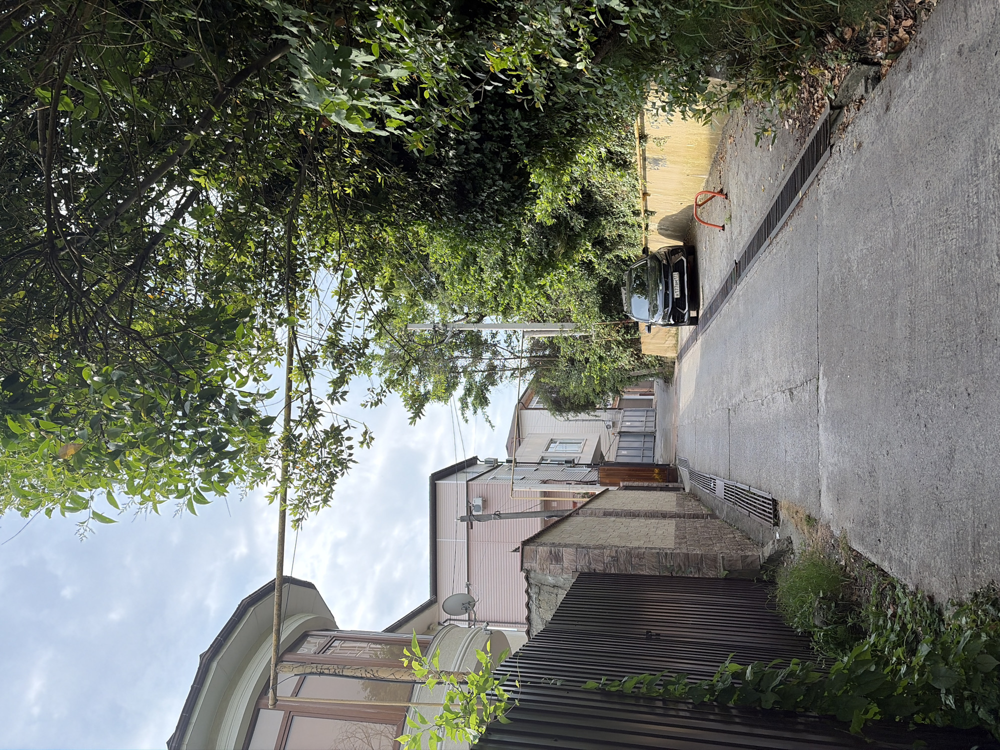 This is a photograph of a narrow, paved alleyway in Sochi, Russia, bordered by buildings and dense greenery. On the left, a modern building with a curved bay window and a dark slatted fence is visible, with a vine climbing the fence. Further down the alley, more buildings with pastel siding and satellite dishes can be seen. On the right, a wall covered in thick, green foliage lines the alley. A black car is parked on the right side of the alley, and an orange metal barrier is situated in front of it. The sky above is cloudy.
