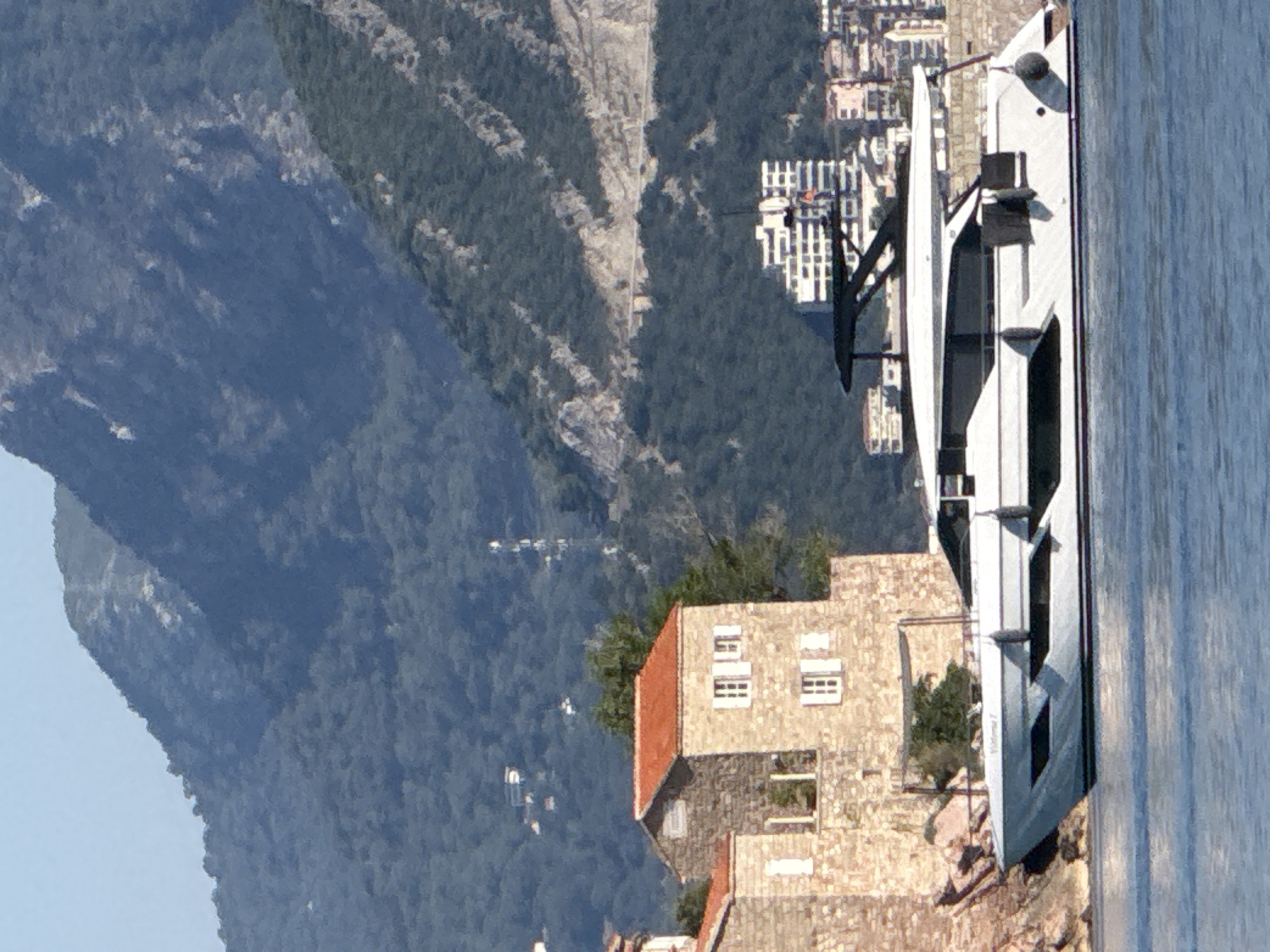 A white yacht with a black frame is docked on the water, with a stone building with a red roof in the background. The building appears to be part of an older structure, possibly a historic monument, and is situated on a rocky outcrop. In the distance, a modern city with tall buildings is visible on the coastline, nestled at the base of a lush green mountain range. The water is calm and reflects the blue sky. The scene is likely located in or near Pržno, Montenegro, given the coastal landscape and architecture.