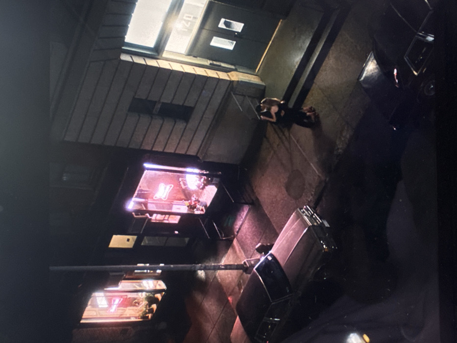 An overhead shot reveals a city street at night. A woman in a black dress lies on the sidewalk near a building entrance marked "140". Adjacent to her, two dark vehicles are parked along the curb. The street features a lighted storefront with an illuminated "N" sign. A lamppost stretches across the scene, and reflections from storefronts can be seen on the sidewalk and the cars.