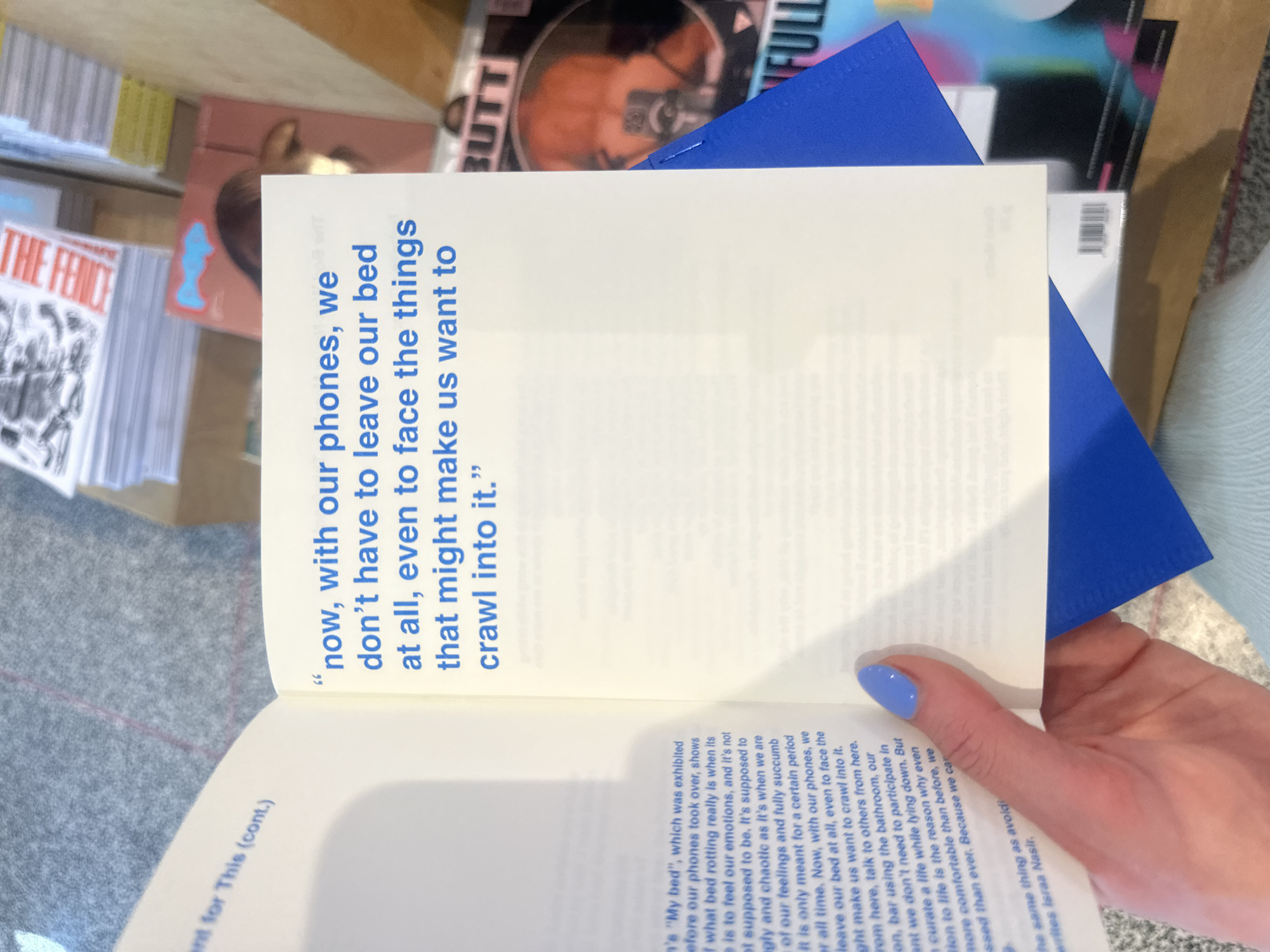 The image shows an open book with text in blue ink. The text reads: "now, with our phones, we don't have to leave our bed at all, even to face the things that might make us want to crawl into it.". A hand with blue nail polish is holding the book open. Other books and magazines, including one titled "THE FENICE" and another partially visible with the word "BUTT" on it, are visible in the background. A blue envelope is also visible. The scene is an interior, possibly a bookstore or a personal collection.