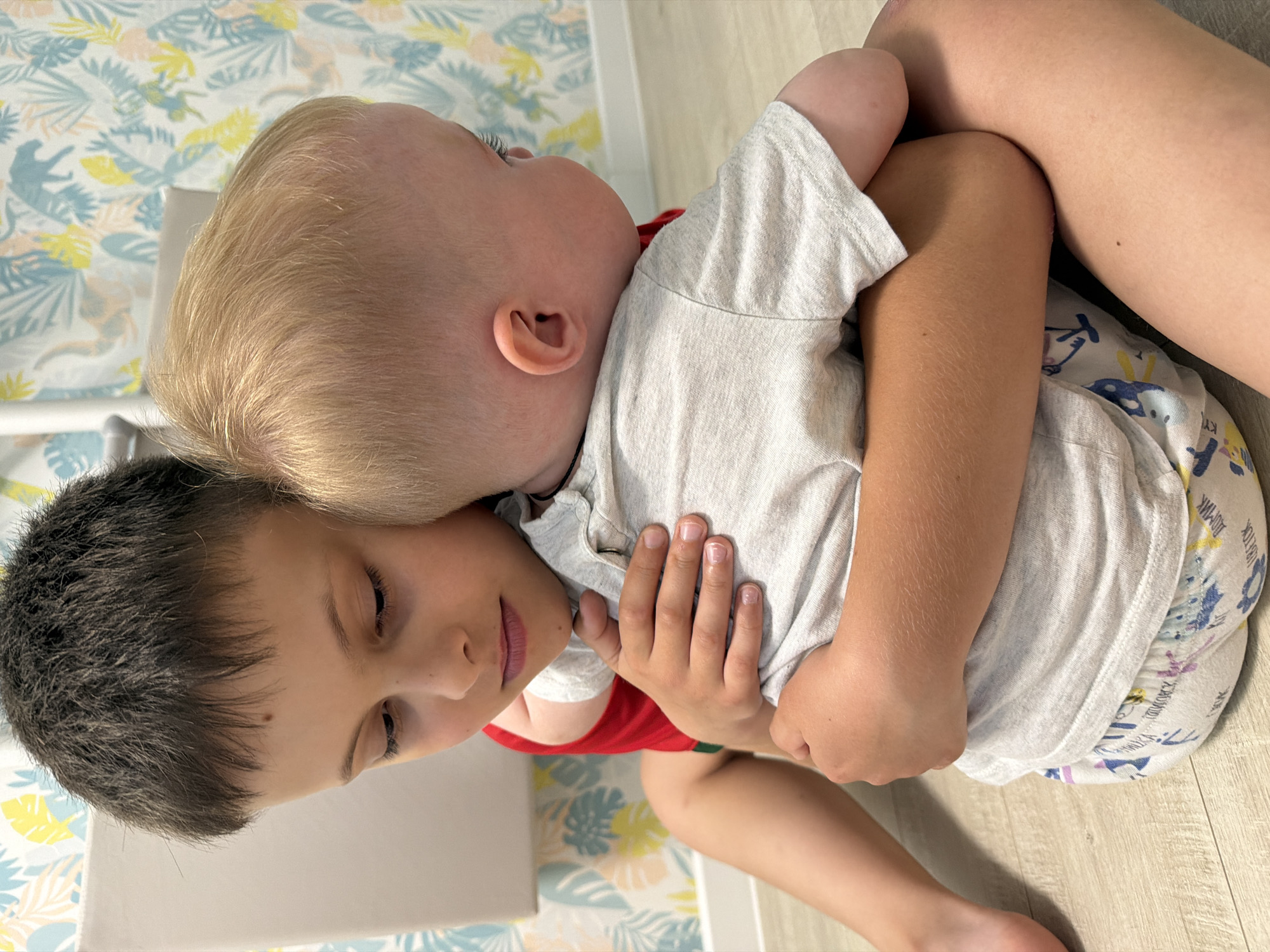 This image captures a tender moment between two children indoors, likely siblings, in what appears to be a home setting in Votkinsk, Russia. An older child with short, dark hair is being affectionately embraced by a younger child with fair, blonde hair. Both children are dressed in casual attire; the older child wears a red undershirt beneath a light grey t-shirt, while the younger child wears a light grey t-shirt and patterned shorts or underwear. The younger child's head rests on the older one's shoulder as their arms are wrapped around, conveying warmth and comfort. The older child looks downwards with a gentle, calm expression, suggesting a reciprocal feeling of affection.
The background features a vibrant wallpaper with light blue, yellow, and beige tropical motifs, including leaves and subtle outlines of jungle animals, typical of a child's room or play area. The floor is light-colored, resembling wooden planks. A white object, possibly a piece of furniture, is partially visible behind the older child's head.
Notable details include the cartoon animal designs and Cyrillic text on the younger child's shorts, with discernible words like "МУМРИК" (MUMRIK) and "ХОХОТОК" (KHOKHOTOK). The older child's fingernails appear well-groomed. The scene is brightly lit, suggesting daytime. The overall mood is one of peace, affection, and familial connection.