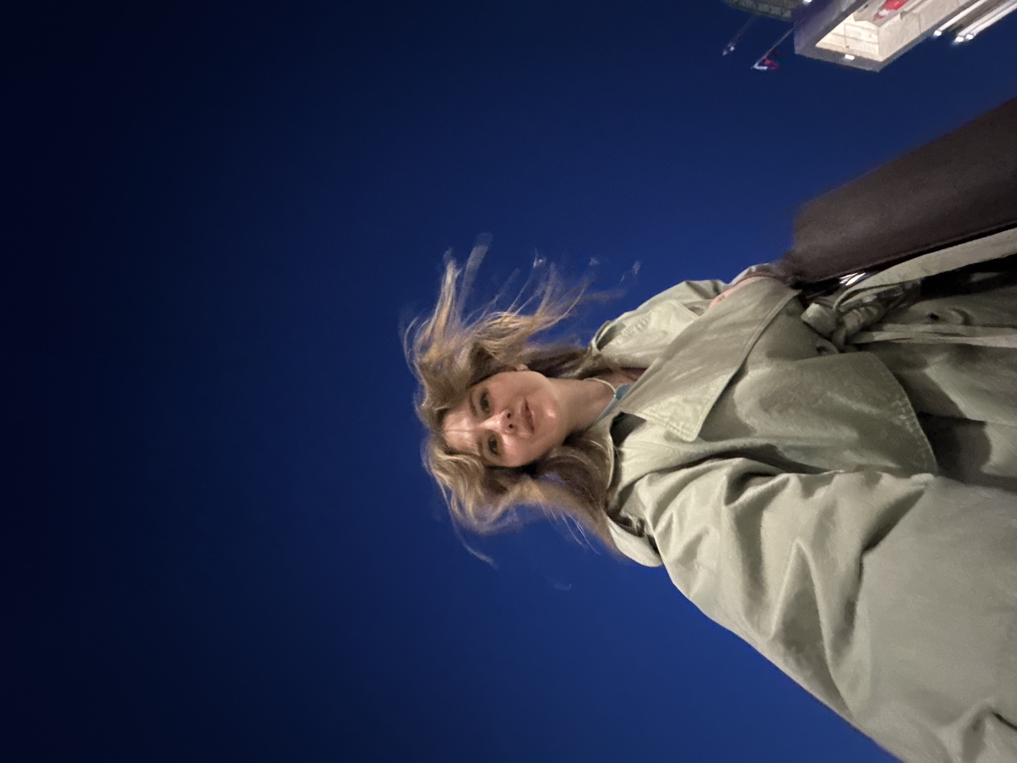 A low-angle portrait shot of a light-skinned female with light brown hair, wearing a light green belted coat, presumably in Perm, Russia. In the upper portion of the image, a building in the distance with red and white architectural elements is barely visible against the dark blue sky. A portion of a dark-colored bag or purse is visible. The woman is looking downward into the camera. The scene appears to be taken at night. The subject's hair is tousled as if caught in a breeze. The background consists of a deep blue color, likely the night sky.