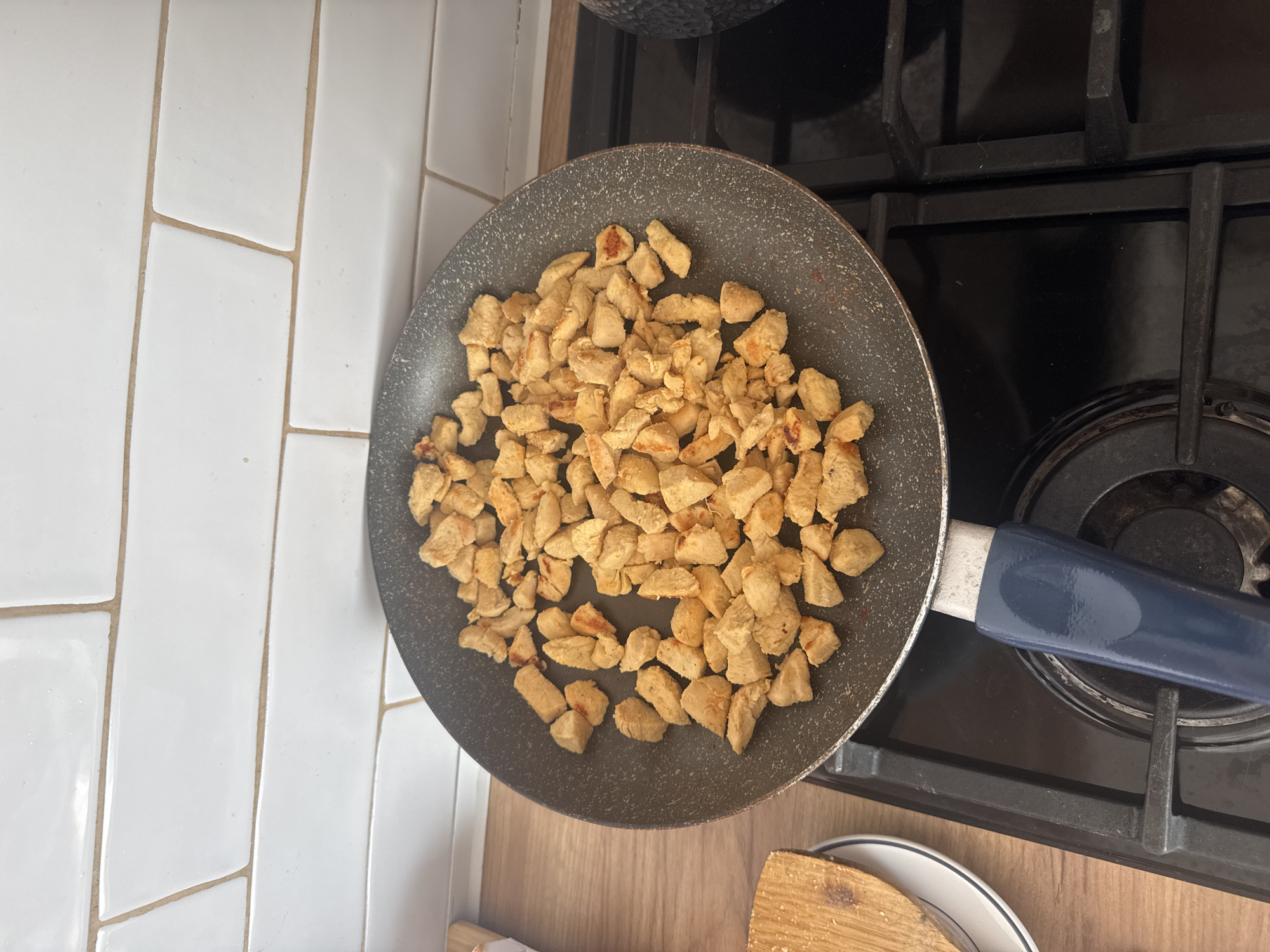 This media file is a close-up, overhead shot of a frying pan filled with diced chicken pieces cooking on a stovetop. The pan is dark gray with a speckled texture and has a blue handle. The chicken is cut into small, bite-sized pieces and appears to be lightly browned, indicating it is being fried or sautéed. The stovetop is black and appears to be a gas stove with grates. In the background, there is a white tiled wall with beige grout lines. The overall setting suggests a kitchen environment. There are no people visible in the frame, and the focus is entirely on the food and cooking apparatus. The lighting appears to be indoor, with no specific indicators of the time of day or weather conditions. No text is visible in the image. The activity depicted is the cooking of chicken.