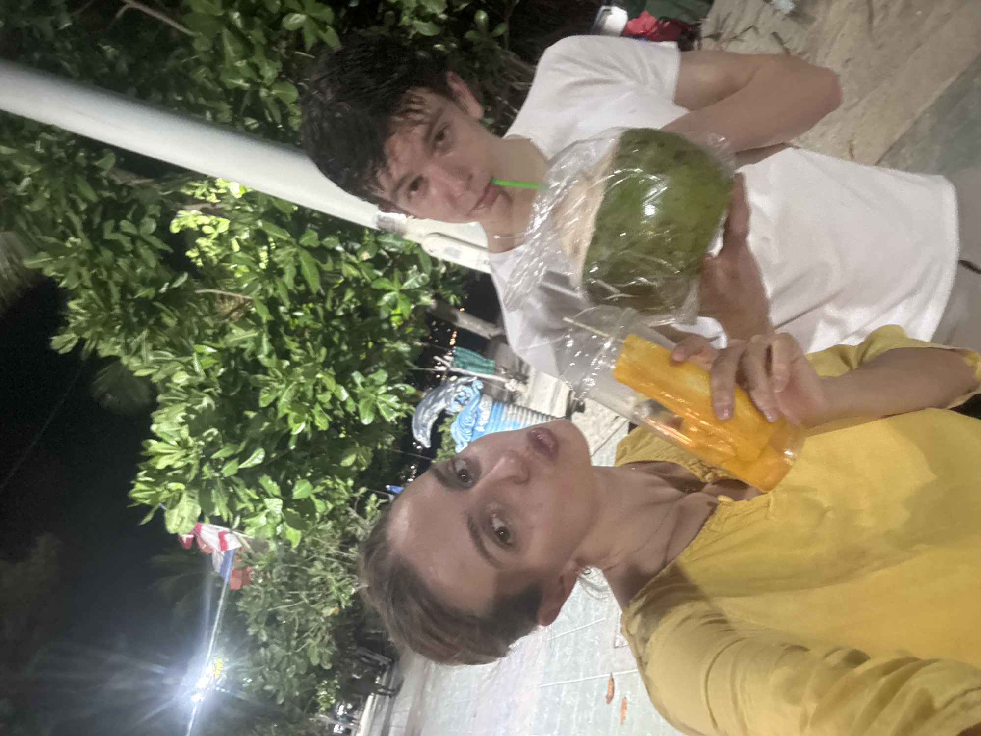 Two individuals are captured in a photograph taken outdoors in what appears to be an urban environment at night. One person is wearing a yellow top and holding a plastic cup containing an orange-colored drink. The other person, slightly out of focus, is wearing a white t-shirt and holds a coconut wrapped in plastic with a green straw inserted. Lush green foliage, possibly trees and bushes, is visible in the background. A light pole with an overhead light is positioned near the foliage. A small Thai flag is partly visible among the foliage. The ground appears to be tiled. The scene is presumably located in the city of Patong, Thailand.