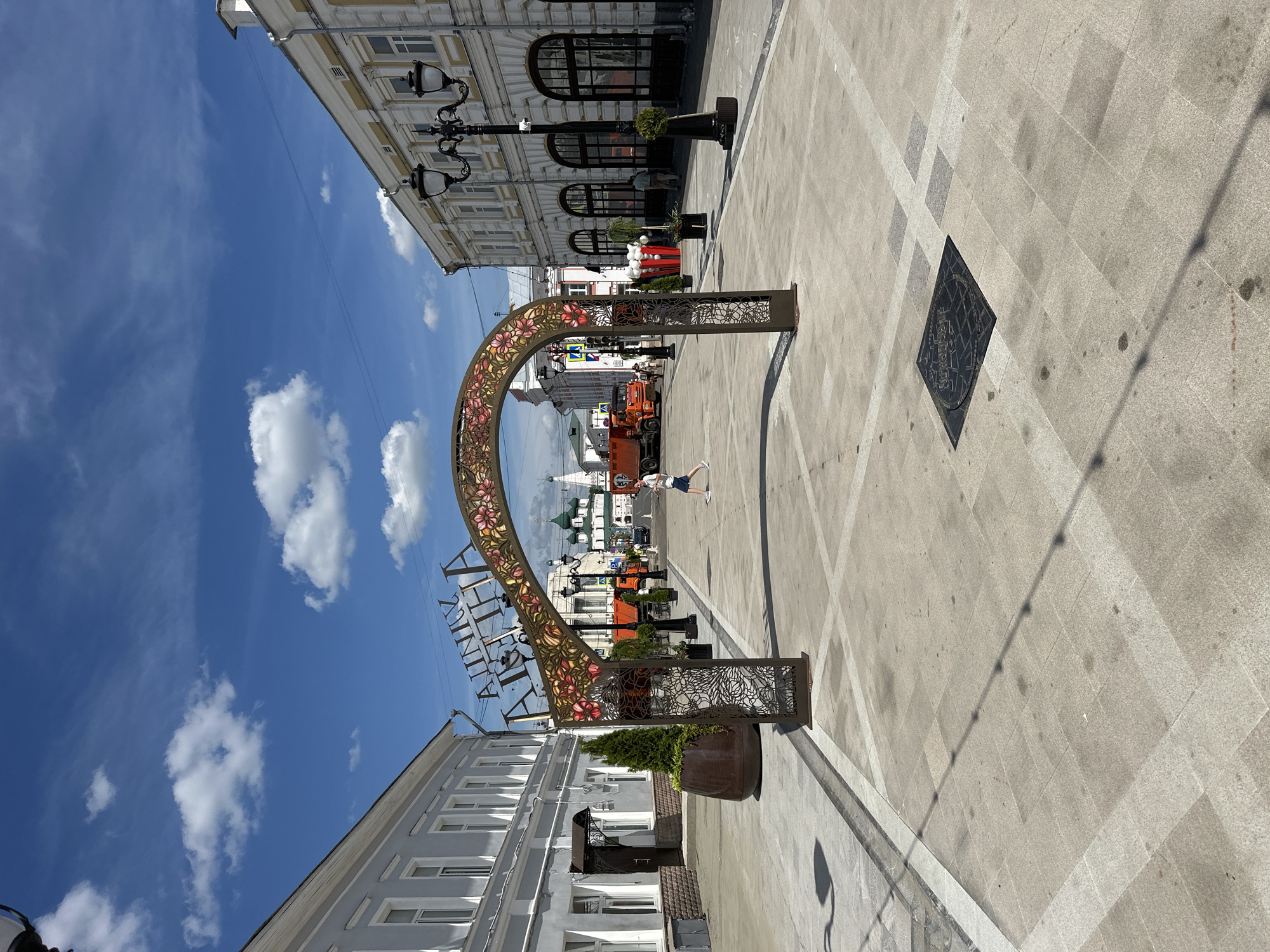 The image depicts a street scene in Putyatino (Semivragovskaya S/A), Russia, on a sunny day. The sky is bright blue with scattered white clouds, indicating clear weather. The perspective is a low-angle shot, looking upwards towards buildings and the sky.
The primary subject is an ornate archway adorned with floral patterns, possibly an entrance to a pedestrian zone or a decorative landmark. The arch is metallic and appears to be a prominent fixture in the urban landscape. In the background, traditional Russian architecture with multiple windows and classical detailing lines the street. Lampposts with multiple globes are also visible, suggesting a historic or well-maintained urban area.
A person is seen in the distance, appearing to walk or pose near the arch. Further down the street, a construction or maintenance vehicle, possibly an orange truck, is parked. Planters with greenery are placed along the sidewalk, adding to the street's aesthetic. The ground is paved with large, light-colored tiles arranged in a patterned grid, with some darker square inlays and a diamond-shaped metal grate visible.
The time of day appears to be midday, given the strong sunlight casting distinct shadows. The overall mood is bright and cheerful, characteristic of a pleasant day outdoors in a city. The visible text on the arch appears to be in Cyrillic script, but it is partially obscured and difficult to read clearly. The scene captures a moment of urban life, with elements of architecture, public art, and everyday activity.