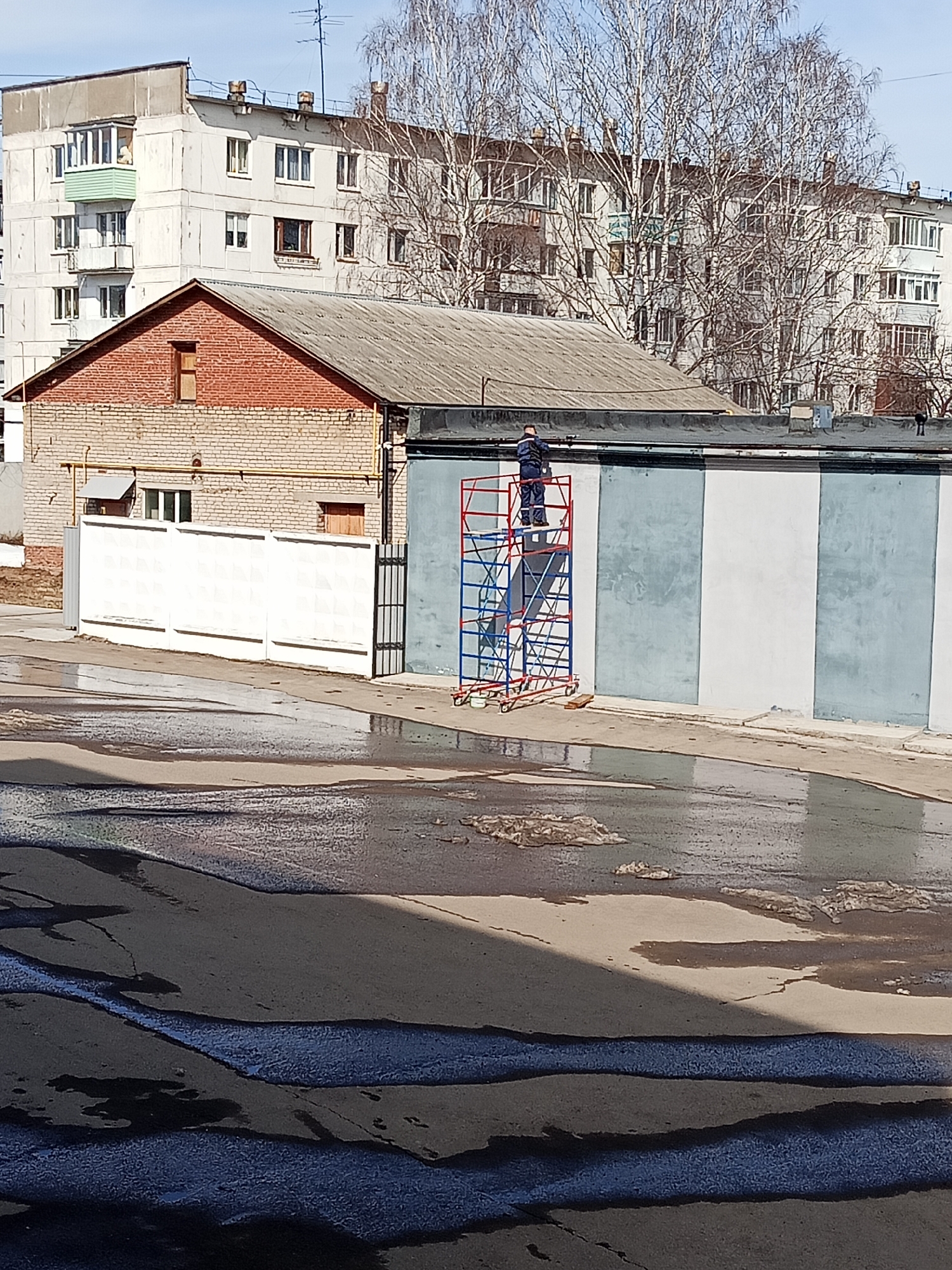 A street scene in the city of Chaikovsky, Russia. A man in blue work clothes stands on a blue and red metal scaffold in front of a gray and white striped concrete wall. To the left is a one-story brick building with a brown roof and a white fence. In the background are several multi-story apartment buildings and bare trees. The foreground is a wet paved surface with puddles.
