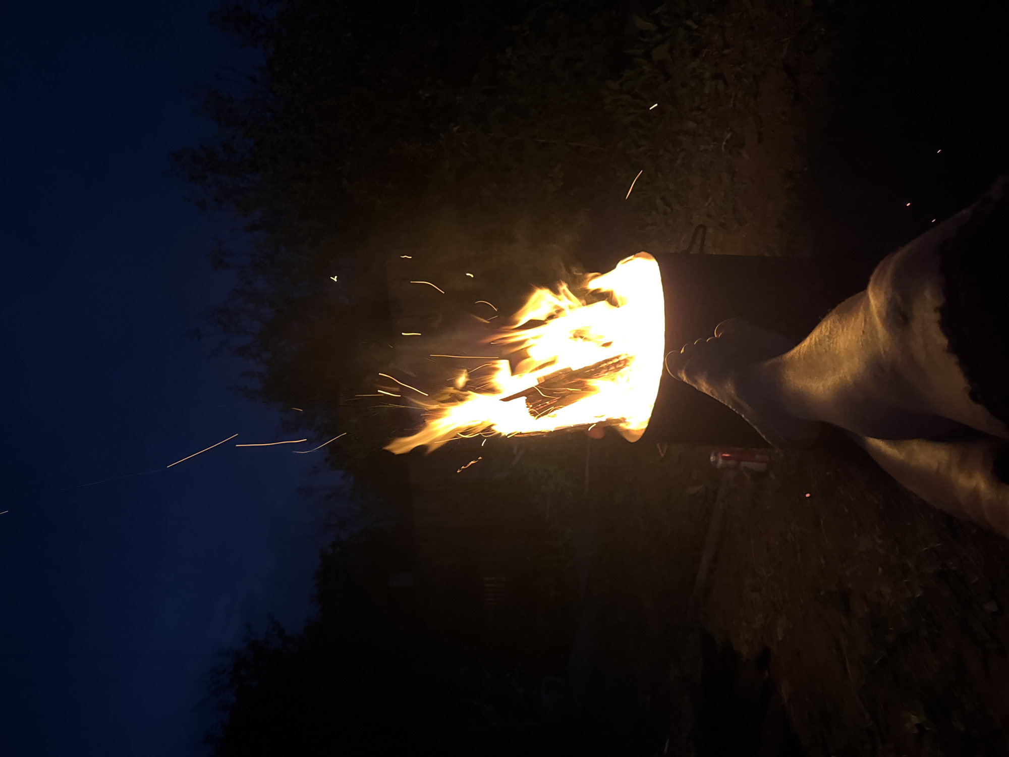 The image depicts a nighttime scene with a bonfire in the foreground. The fire is burning brightly in a metal drum, casting a warm glow on the surroundings. Sparks and embers are rising from the flames, some of which are visible as streaks of light against the dark sky. In the lower right portion of the image, a person's legs are visible, indicating they are sitting or lying down near the fire. The background consists of dark foliage and trees, suggesting an outdoor setting, possibly a backyard or a park. The sky is a deep blue, characteristic of twilight or early night. There is no specific information within the image to indicate the location in Chaikovsky, Russia.