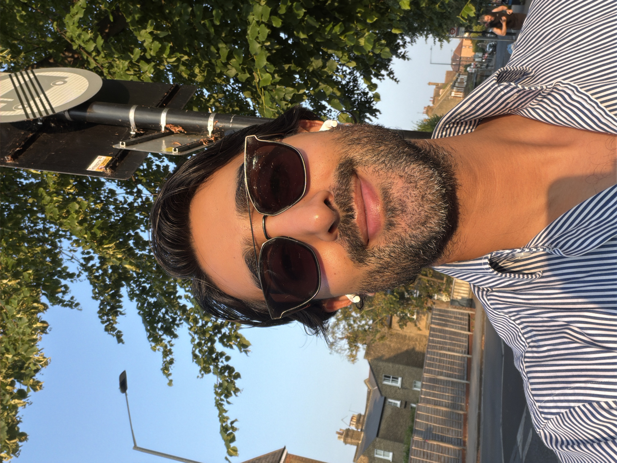 A man with dark hair, a beard, and sunglasses is taking a selfie outdoors on a street in London, United Kingdom. He is wearing a blue and white striped shirt. The background shows a tree, a street lamp, a traffic sign, and residential buildings. The sun is shining, casting a warm glow on the man's face. The man appears to be looking at the camera and smiling slightly. In the distance, another person is visible jogging down the street. The street is paved and there are cars parked along the side. A wooden fence is also visible in the background.