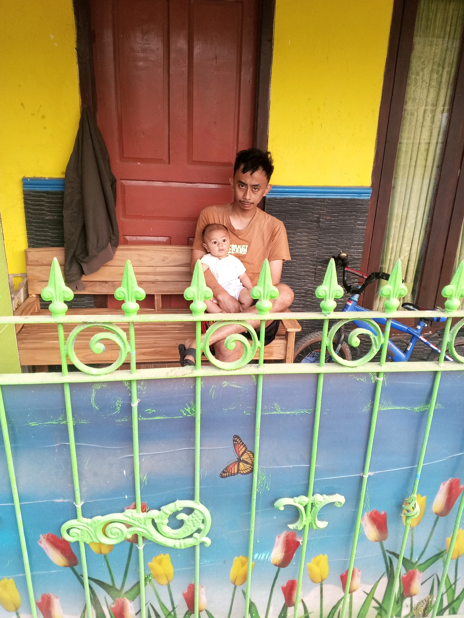 A young man is sitting on a wooden bench on a porch, holding a baby in his arms. He is wearing an orange t-shirt and black shorts. The baby is wearing a white onesie and is looking to the right. Behind the bench, there is a dark brown wooden door and a yellow wall. A dark brown jacket is hanging on the left side of the door. To the right of the bench, there is a blue bicycle with black handlebars and a red bell. The porch is partially obscured by a green metal fence with a patterned backdrop featuring tulips and a butterfly. The fence has decorative scrollwork. The setting appears to be a residential area.
