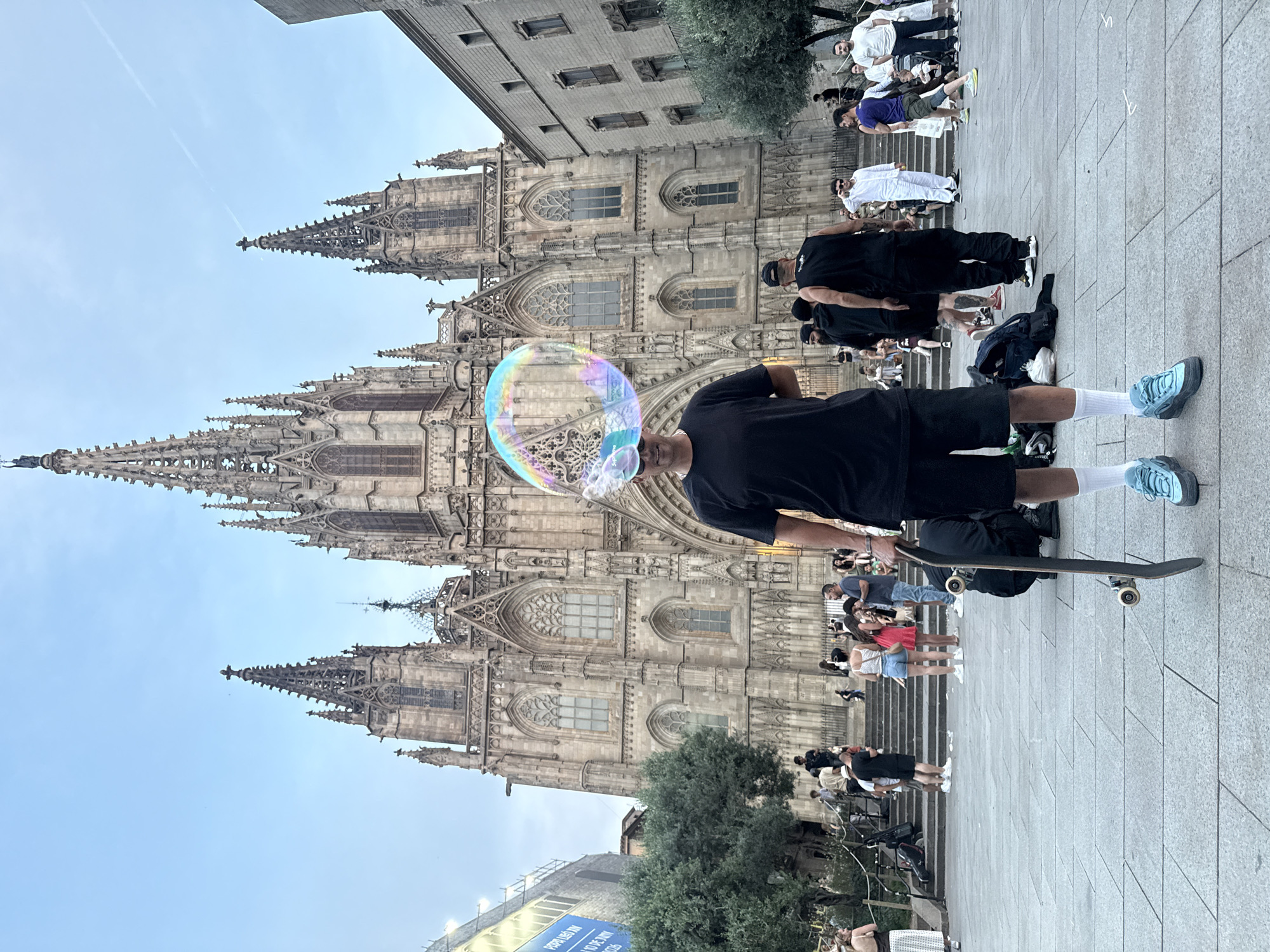 A man stands on a stone-paved area, holding a skateboard, with a large soap bubble floating in front of his face. He is wearing a black t-shirt, black shorts, white socks, and blue sneakers. The Cathedral of Barcelona, a Gothic architectural structure with spires, towers behind him. There are other people present, some standing on the steps of the Cathedral. The sky is blue and clear. A sign is at the bottom of the image. The photo appears to be taken in a public square in Barcelona, Spain.