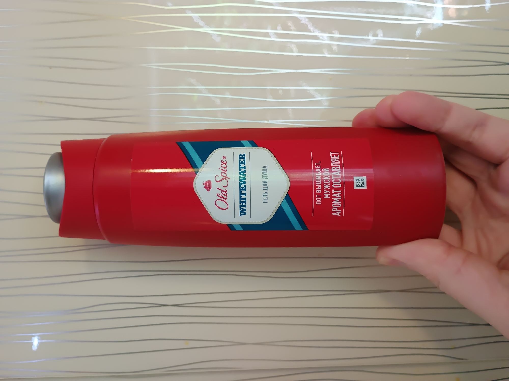 A red Old Spice Whitewater body wash bottle is held in a person's left hand. The bottle has a silver cap and a label with text in Russian. The label reads "Old Spice", "WHITEWATER", and "ГЕЛЬ ДЛЯ ДУША" (Gel for shower). Below this, it says "ПОТ ВЫШИБАЕТ, МУЖСКОЙ АРОМАТ ОСТАВЛЯЕТ" (Sweat drives away, masculine aroma remains). A QR code is visible on the lower part of the label. The background is a wall with vertical cream-colored lines. The location of this image is not provided in the description.