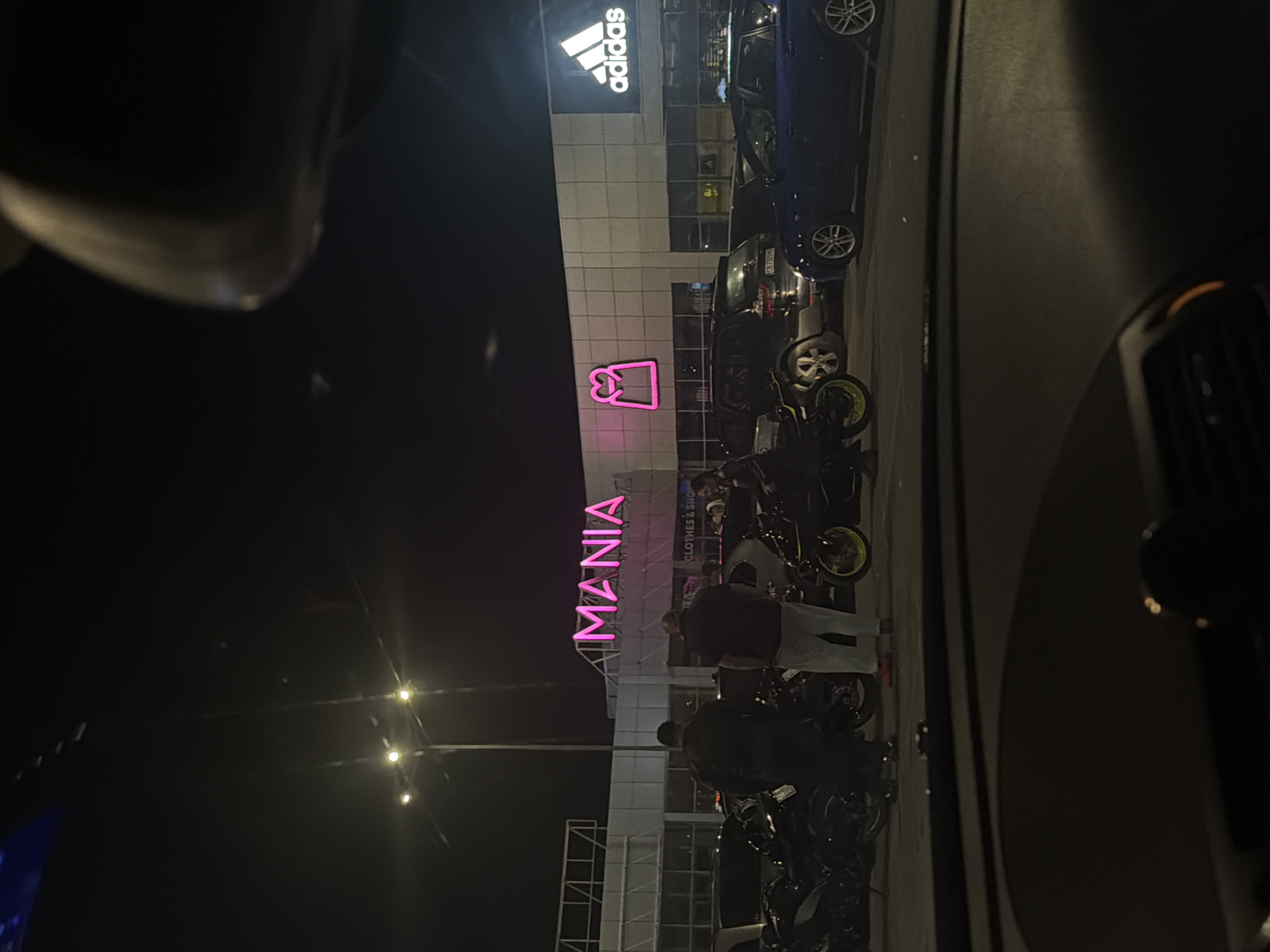 At night in Sofia, Bulgaria, a view from inside a car reveals an Adidas store adjacent to a Mania retail location, the latter with its name illuminated in bright pink neon letters. Several motorcycles and a couple of parked cars are visible in front of the building. People are present near the front of the building. Streetlights are seen in the background. The image is framed by elements of the car's interior.