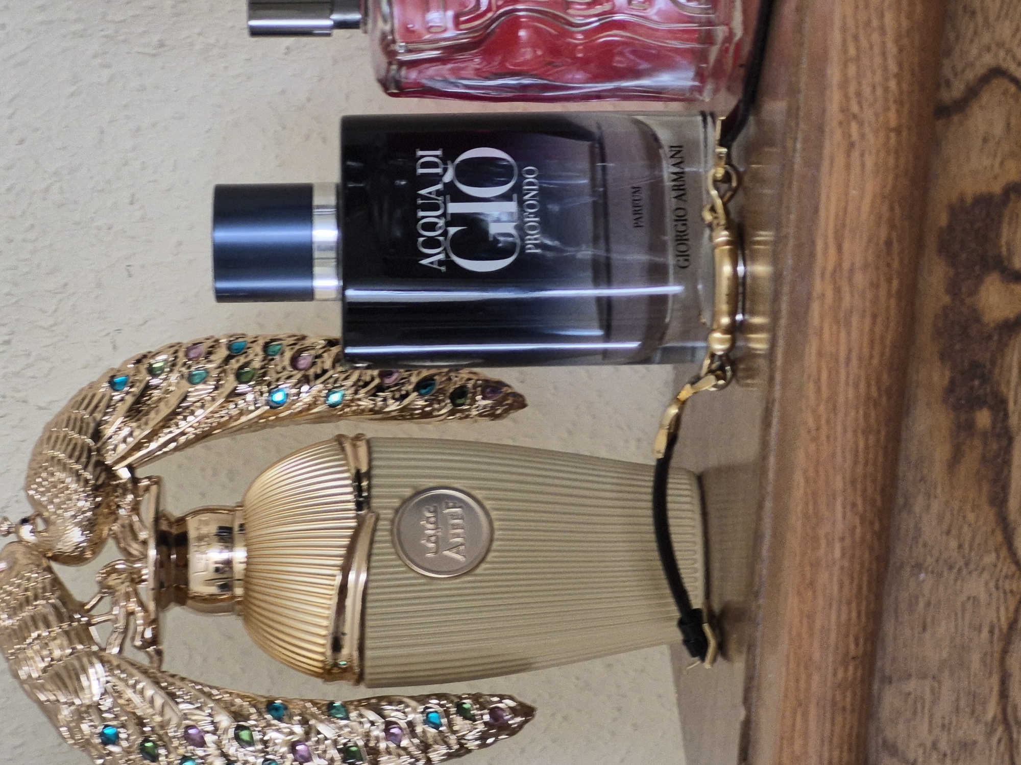 This is a close-up, eye-level, indoor photograph of three perfume bottles on a wooden shelf. The leftmost bottle is gold with a fluted design and decorative gold peacocks with jeweled feathers as a cap. In the center is a dark glass bottle of "ACQUA DI GIO PROFONDO PARFUM" by GIORGIO ARMANI. To the right is a red, ridged glass perfume bottle. A black and gold bracelet or strap is draped across the shelf in front of the bottles. The background is a textured, light-colored wall. The image is well-lit, with natural light coming from the left. There are no people visible.