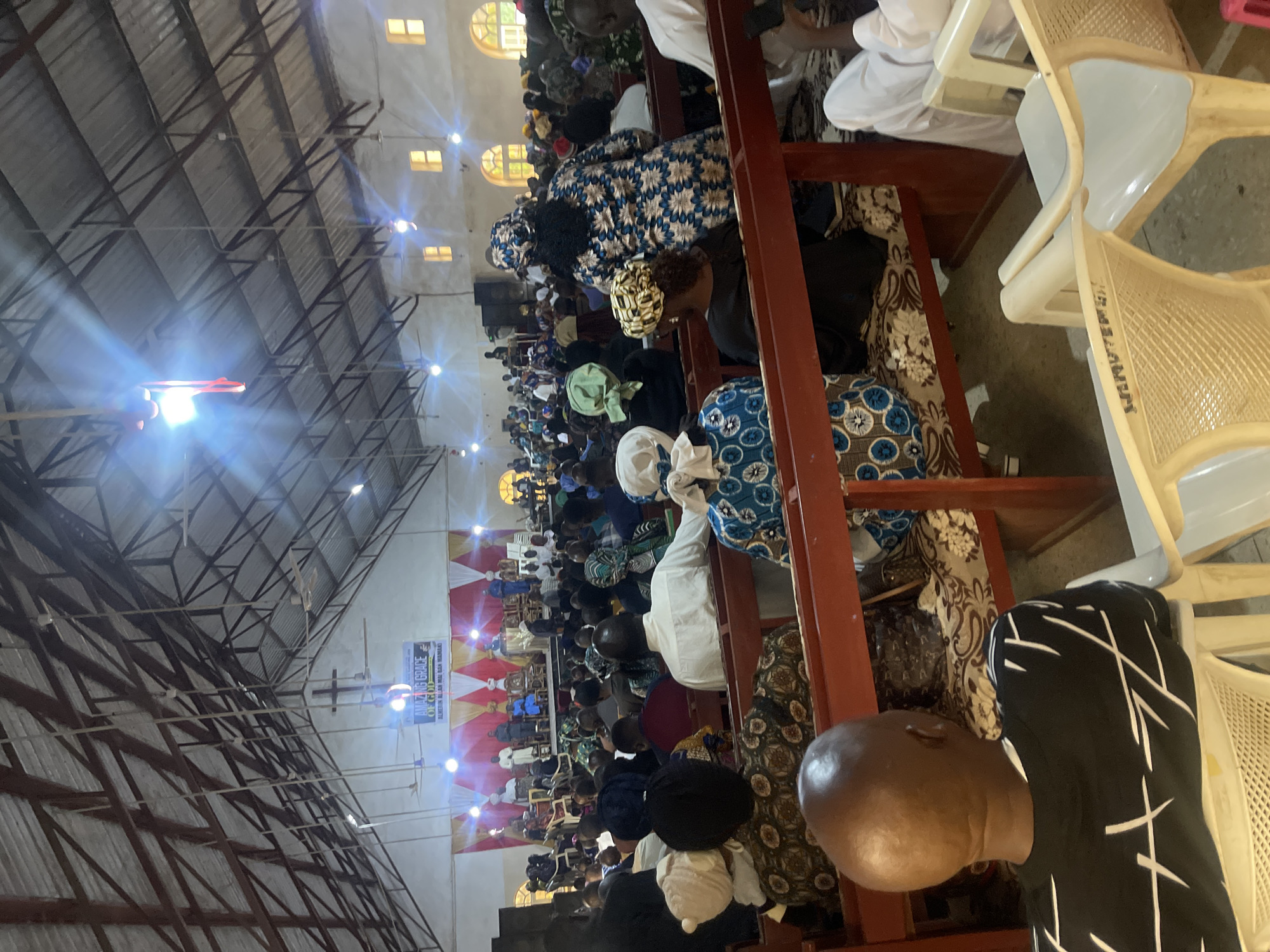 An interior view of a crowded church, likely in Jos, Nigeria, captures a congregation seated on wooden pews and plastic chairs. The church has a high, exposed metal truss roof with numerous hanging lights and ceiling fans. A banner proclaiming "AMAZING GRACE OF GOD" is prominently displayed at the front, above a stage where several individuals are gathered. The congregation, diverse in age and attire, includes men, women, and children, many wearing head coverings and traditional African clothing. The overall atmosphere suggests a religious service or event.