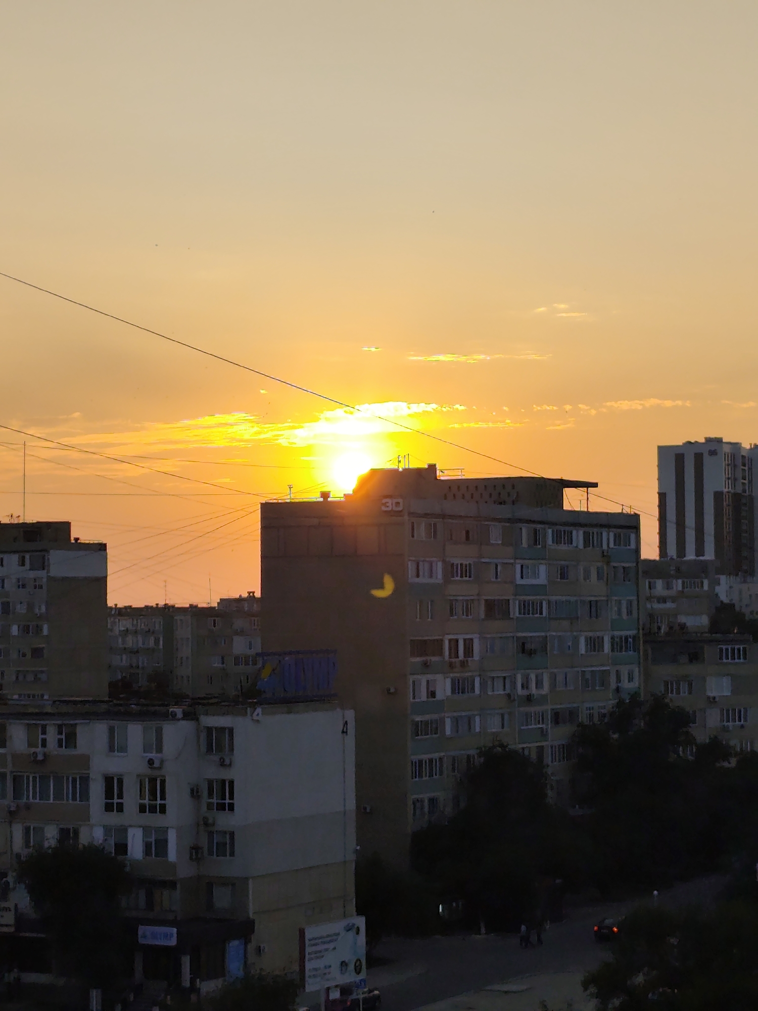 A low-angle, medium shot captures a cityscape at sunset in Aktau, Kazakhstan. The sun, a bright orange orb, is partially obscured by the top edge of a large, multi-story apartment building in the mid-ground, its rays casting a warm, golden glow across the sky and the upper floors of the buildings. The sky itself is a gradient of soft orange and pale yellow, with a few wispy clouds scattered across it.
Several apartment buildings dominate the scene. In the foreground, to the left, is a lighter-colored, multi-story residential building with visible windows and air conditioning units. To the right of this, and slightly behind it, is a taller, darker apartment building with rows of windows. Further back, a very tall, slender modern building with a striped facade rises into the sky. The buildings appear to be typical Soviet-era architecture, common in many cities of Kazakhstan.
Overhead, a complex network of power lines crisscrosses the frame, adding a utilitarian element to the otherwise picturesque sunset.
In the lower part of the image, a darker area suggests the ground level, with trees casting shadows and a road visible. A few indistinct figures are present on the ground, as is a car with its taillights illuminated. A blue dumpster or container is visible on the roof of one of the apartment buildings. The overall impression is one of urban life continuing as the day ends, with the dramatic sunset providing a striking backdrop.