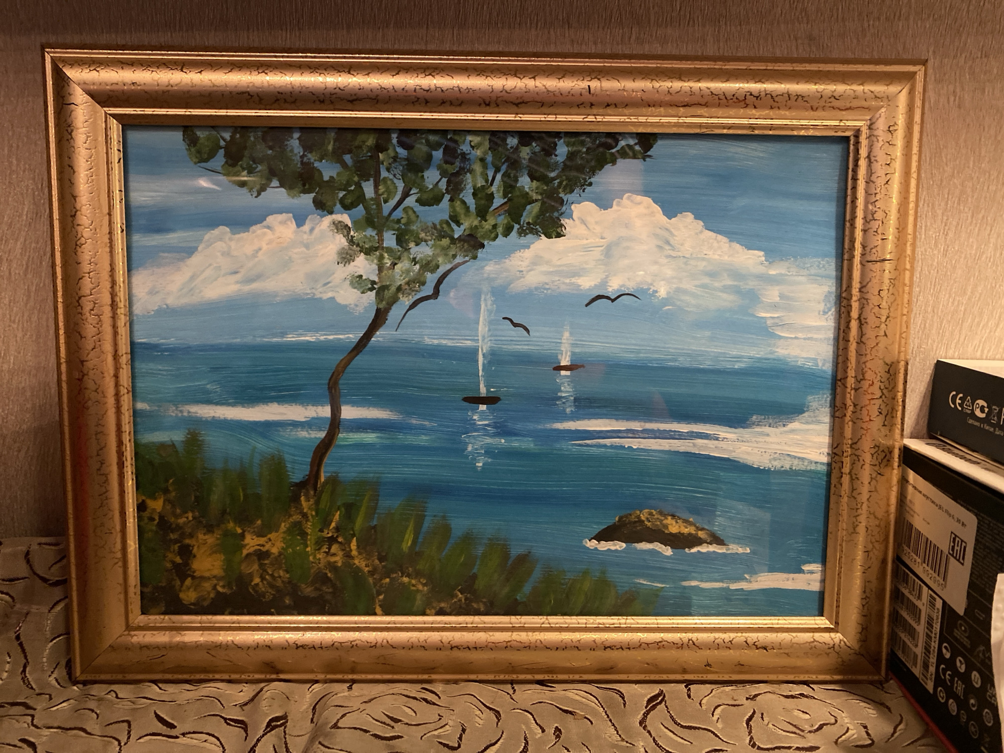The image shows a framed painting of a seascape. The painting depicts a bright blue ocean with white-capped waves under a sky with fluffy white clouds. In the foreground, there is a tree with green leaves on the left side of the canvas, and a rocky island in the distance on the right. Two sailboats with brown hulls are visible on the water. There are also two black birds flying in the sky. The painting is framed in a gold-colored frame with a textured surface. The painting is displayed on a surface covered with a patterned cloth. In the background, to the right of the painting, there is a cardboard box with some Russian text and certifications. The scene is likely indoors, given the lighting and the presence of the box. The location in Ivanteyevka, Russia is not discernible from the image.