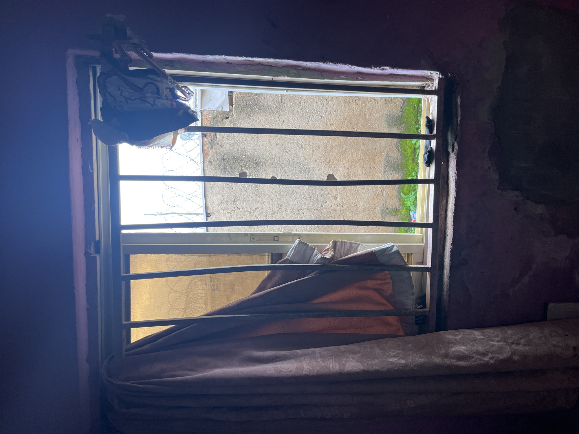 A barred window of a room in Jos, Nigeria, shows a view to the outside. A thick, patterned curtain, predominantly in shades of pink and rust, is drawn to the left side of the window, obscuring a portion of the view. On the right side of the window, items of clothing are draped over the bars, including what appears to be a black jacket or shirt, and other garments with patterned designs.
The window frame is metallic and appears to be made of vertical bars. Outside, a concrete wall with some visible damage and patches of green vegetation can be seen. Barbed wire is also visible on top of the exterior wall, suggesting a measure of security. The interior of the room, where the photograph is taken from, appears dimly lit and the walls are a dark, reddish-pink color with some signs of wear and tear. The windowsill is also visible, with some dark objects resting on it. The overall impression is one of a somewhat modest and perhaps aged living space.