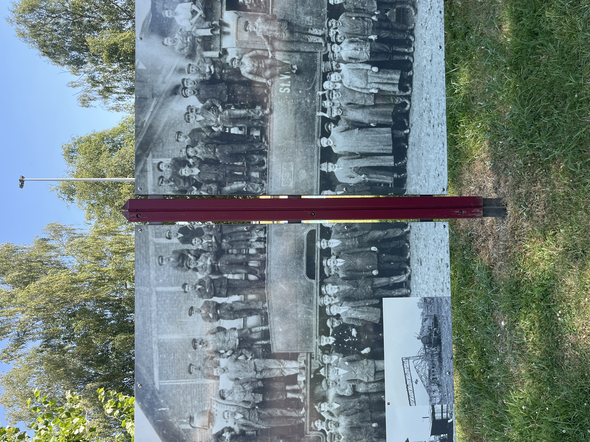 The image is a photograph of a large historical outdoor exhibit in the city of Saint-Vith, Belgium. The exhibit features a black and white panoramic photograph of a group of men, likely workers, standing and sitting on and around a train. They are dressed in work clothes, some with hats. The exhibit is supported by a maroon metal post. In the background, green trees and a bright blue sky are visible. Below the main photographic display, there is a smaller black and white image showing a damaged building and a train, possibly depicting an event in Saint-Vith's history. The exhibit is set against a backdrop of green grass.
