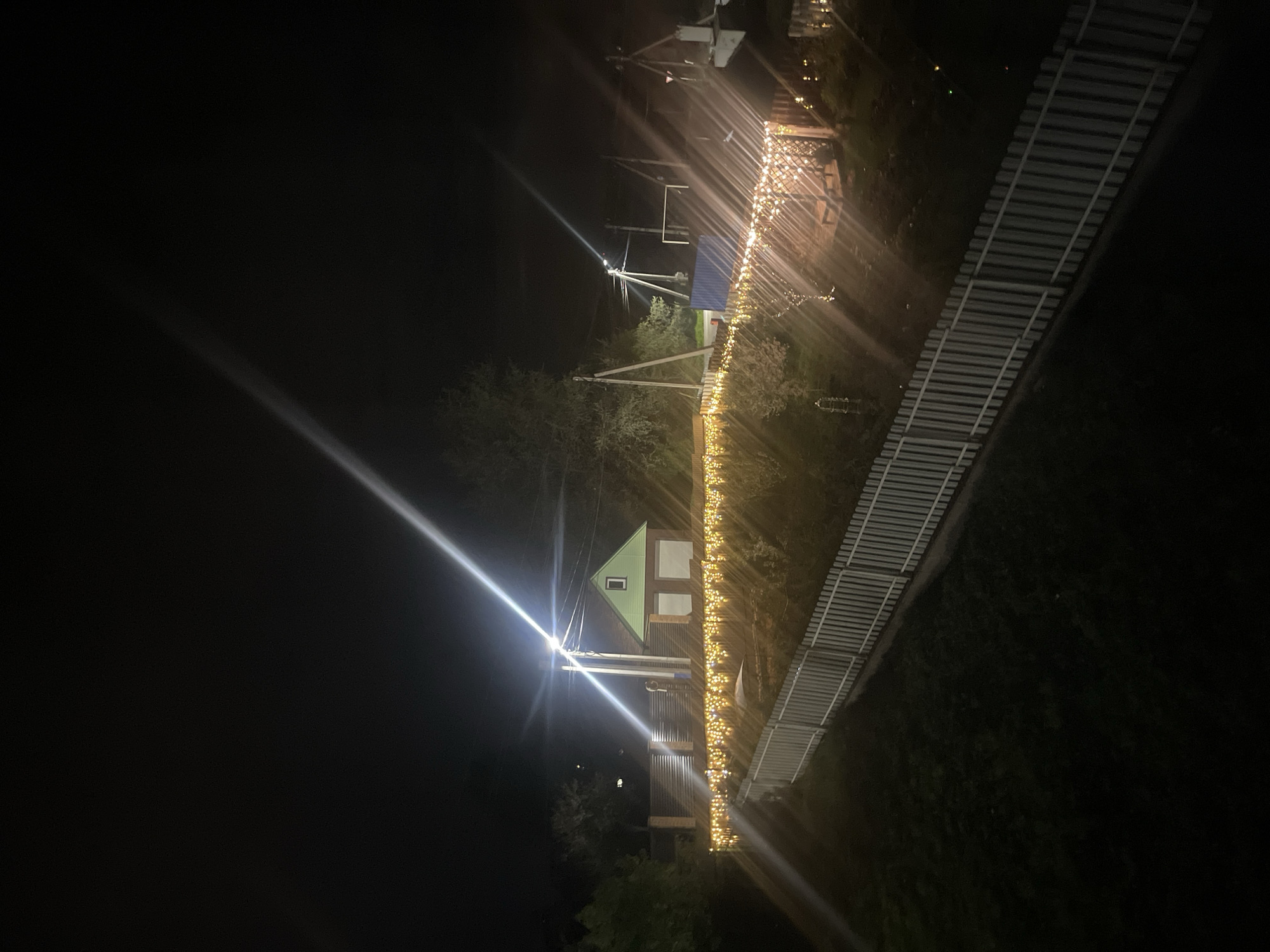 The image depicts an outdoor scene at night in what appears to be the city of Хлябово, Russia. A small house with a green roof is visible, illuminated by a bright light source that causes significant lens flare. A fence adorned with string lights runs alongside the house. In the background, there is a structure with a blue roof supported by wooden posts, also lit by string lights. A slatted bridge or walkway is seen on the right side of the frame. The surroundings appear dark, with only minimal ambient light. The image is rotated counter-clockwise.