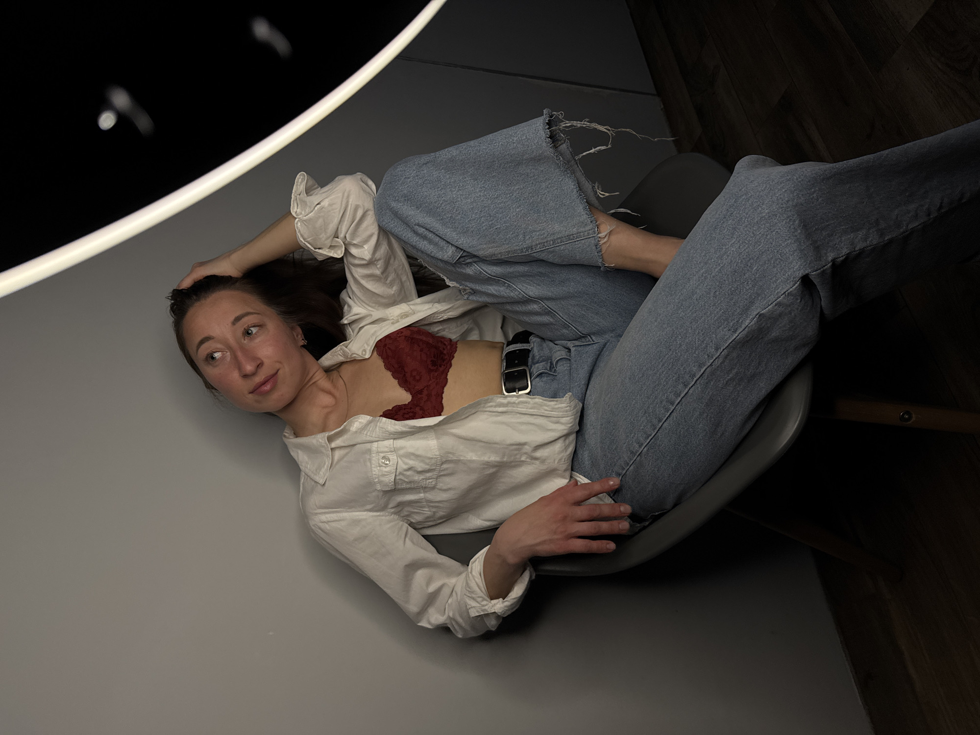 The image shows a woman lying in a chair in an indoor space. The woman has long, dark hair and is wearing a white, unbuttoned shirt, a red bra, and blue jeans with a black belt. She is reclined in the chair, with one hand behind her head and the other resting on the chair. The room has grey walls and a dark ceiling with a circular light fixture. Based on the surrounding images and the source, the location is the city of Chaikovsky, Russia.